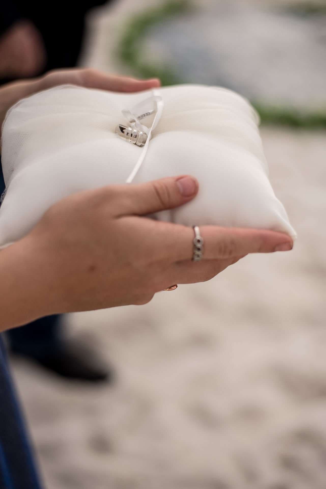 Hands holding a white satin ring pillow with two wedding bands tied on by a ribbon, on the beach