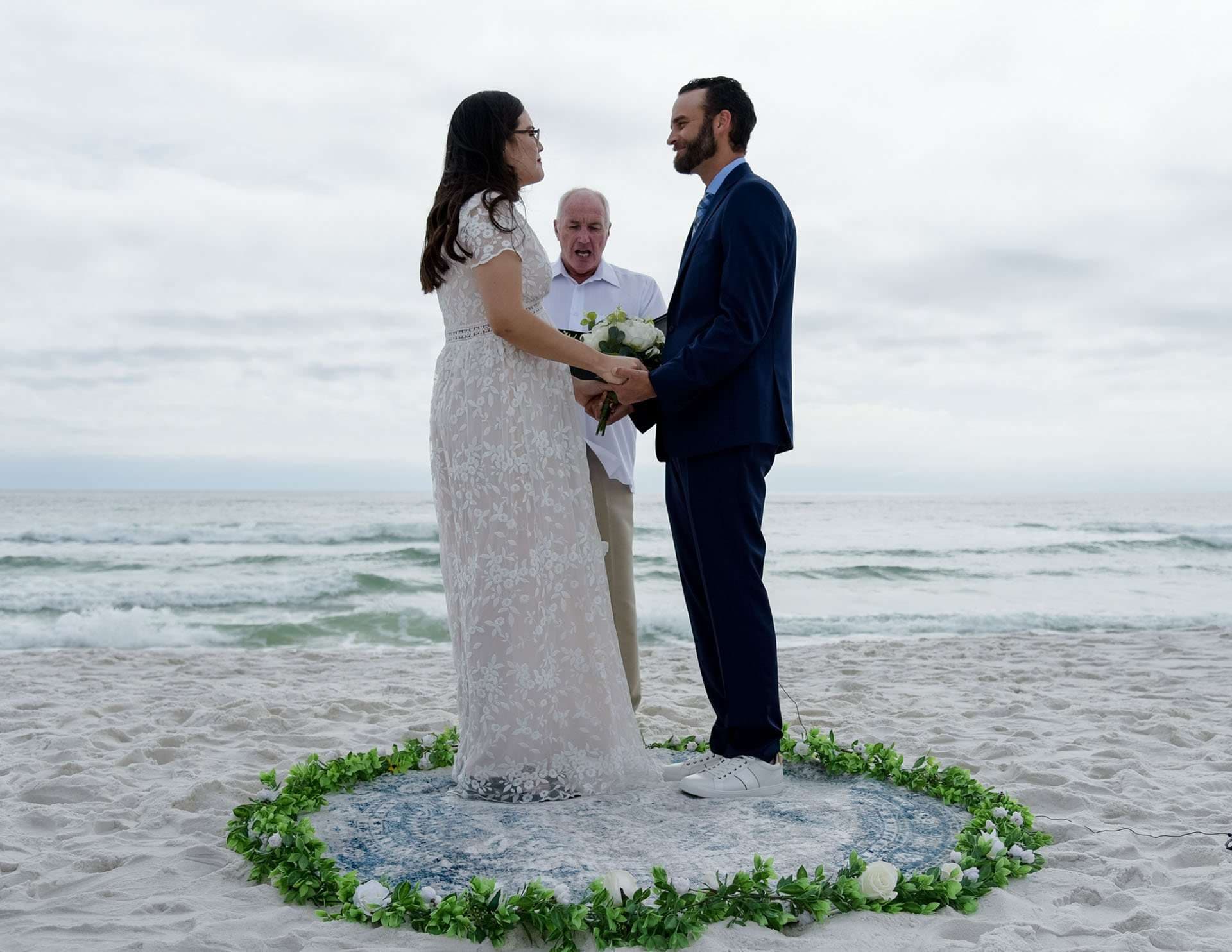Couple exchanging vows on the beach inside a circle of greenery while an officiant stands behind them