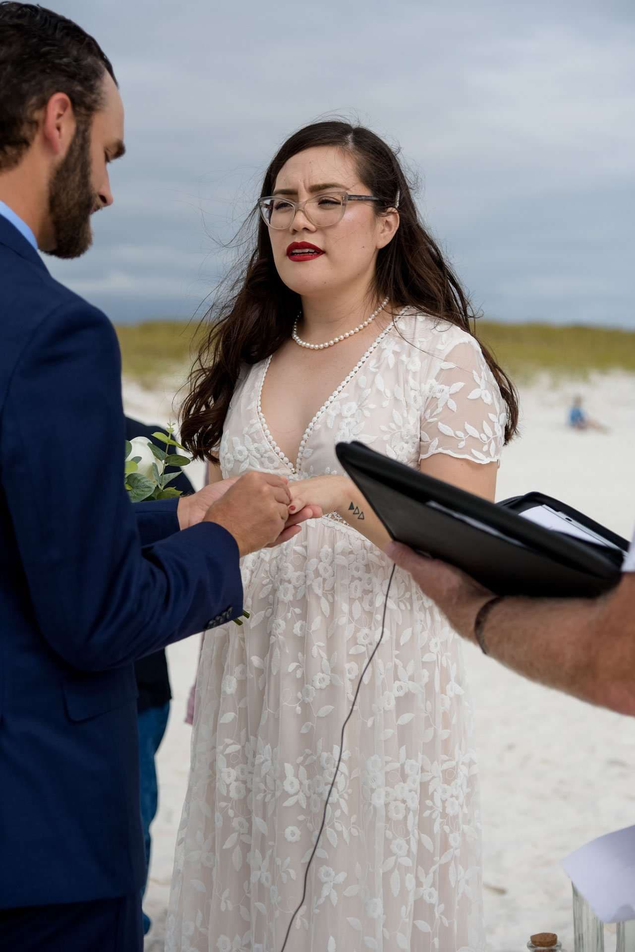 Couple exchanging rings on the beach on Okaloosa Island, bride in white lace dress and glasses, officiant holding a folder