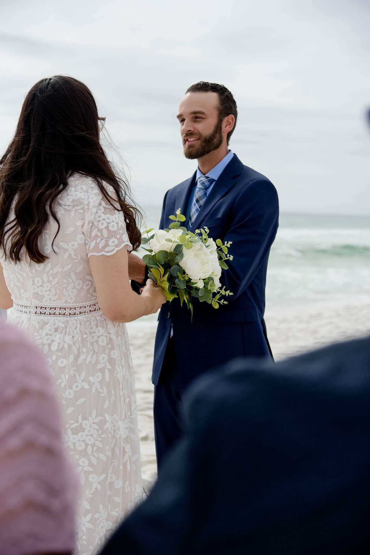 Smiling bearded groom in navy suit holding a white bouquet facing his bride in a lace dress on the beach