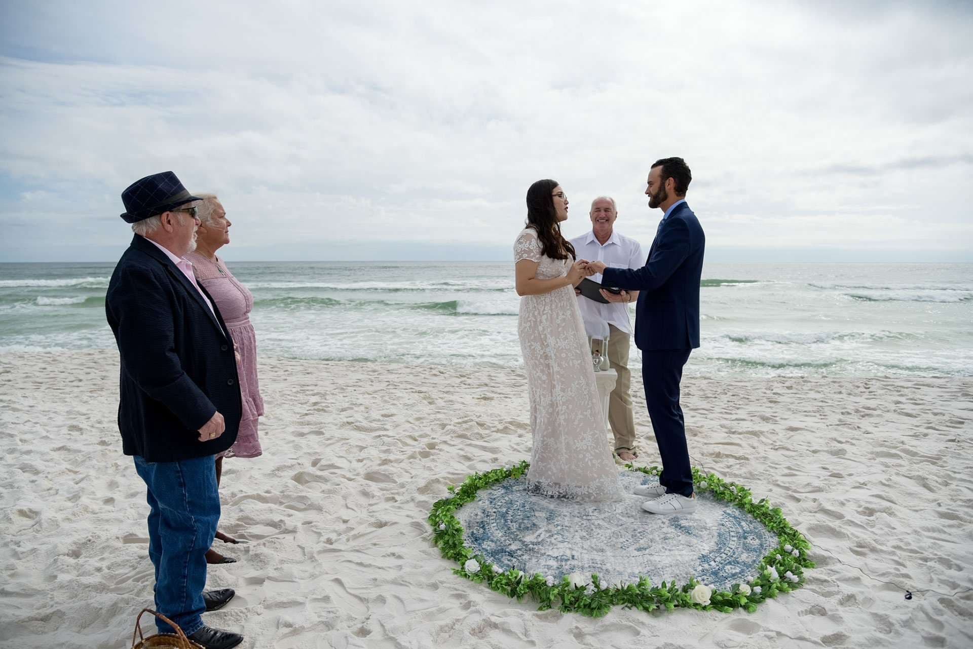 Couple exchanging vows on Okaloosa Island beach inside a circle of greenery, with an officiant and two guests watching