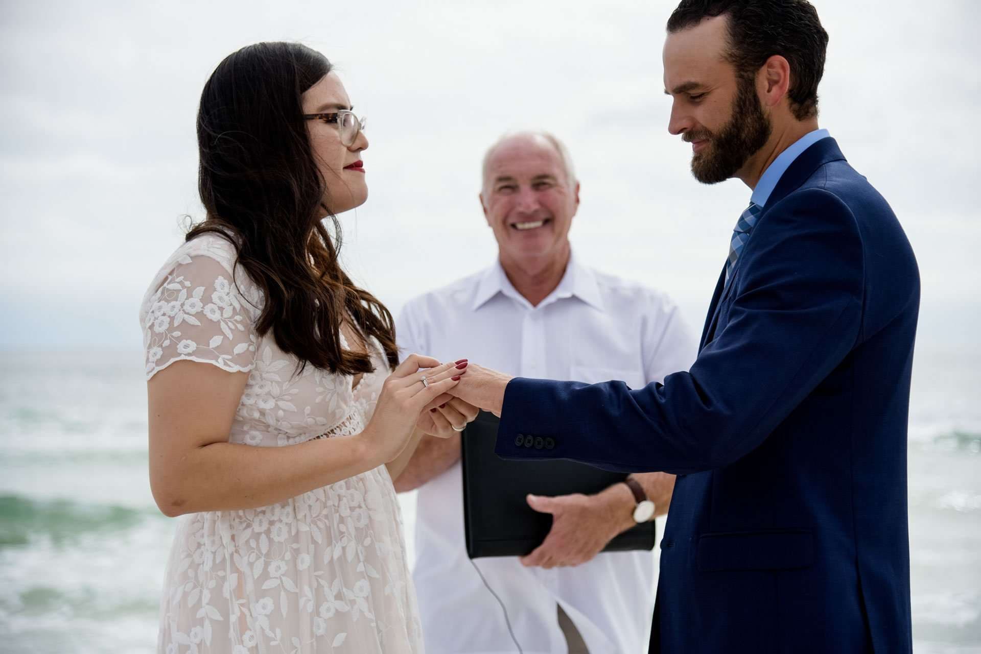 Couple exchanging rings on the beach on Okaloosa Island while a smiling officiant looks on behind them
