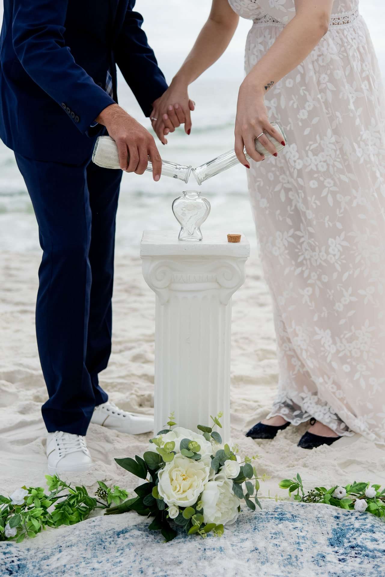 Couple pouring sand from two vials into a glass heart during a beach ceremony, beside a white column and flowers