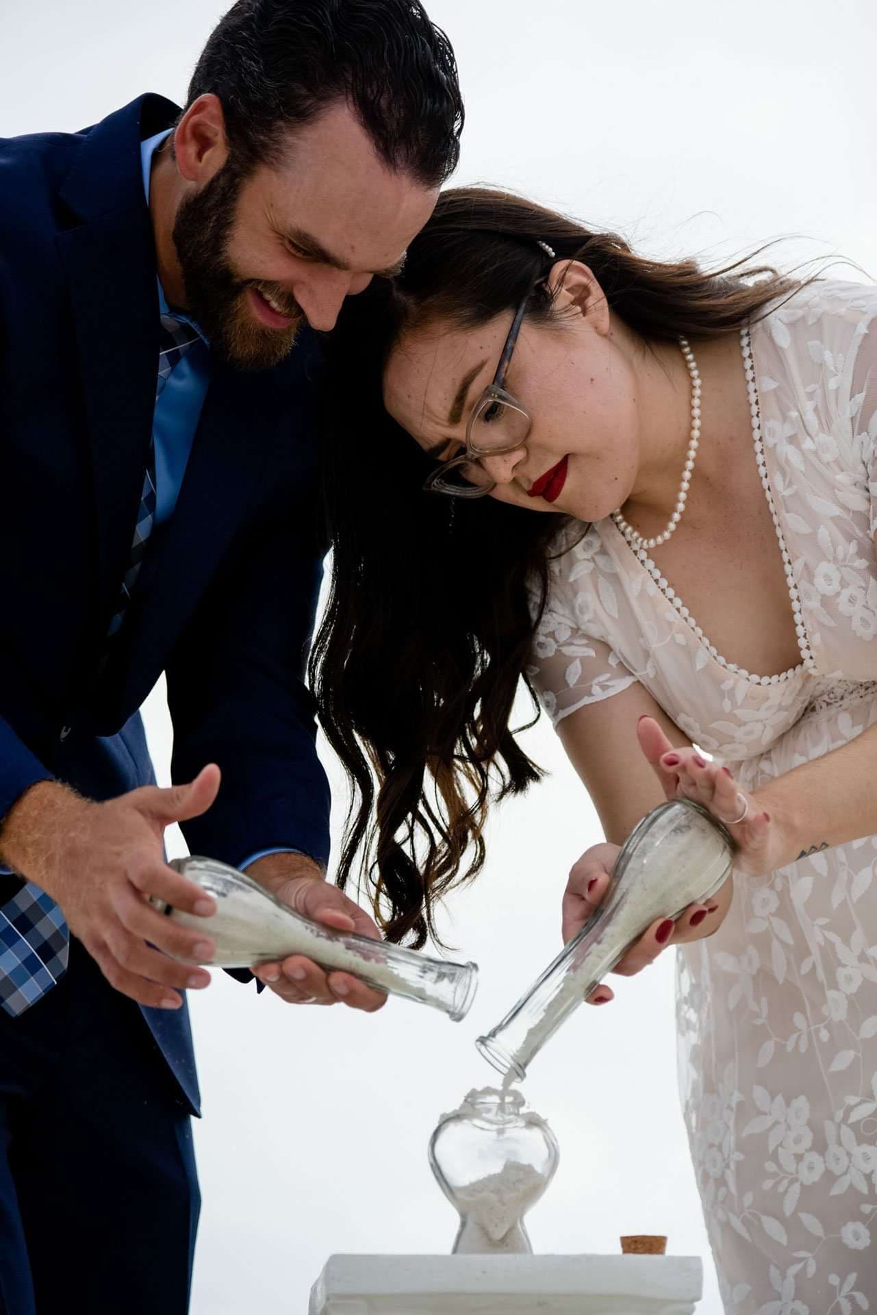 Couple pouring white sand from two glass bottles into a heart-shaped vessel during a wedding sand ceremony