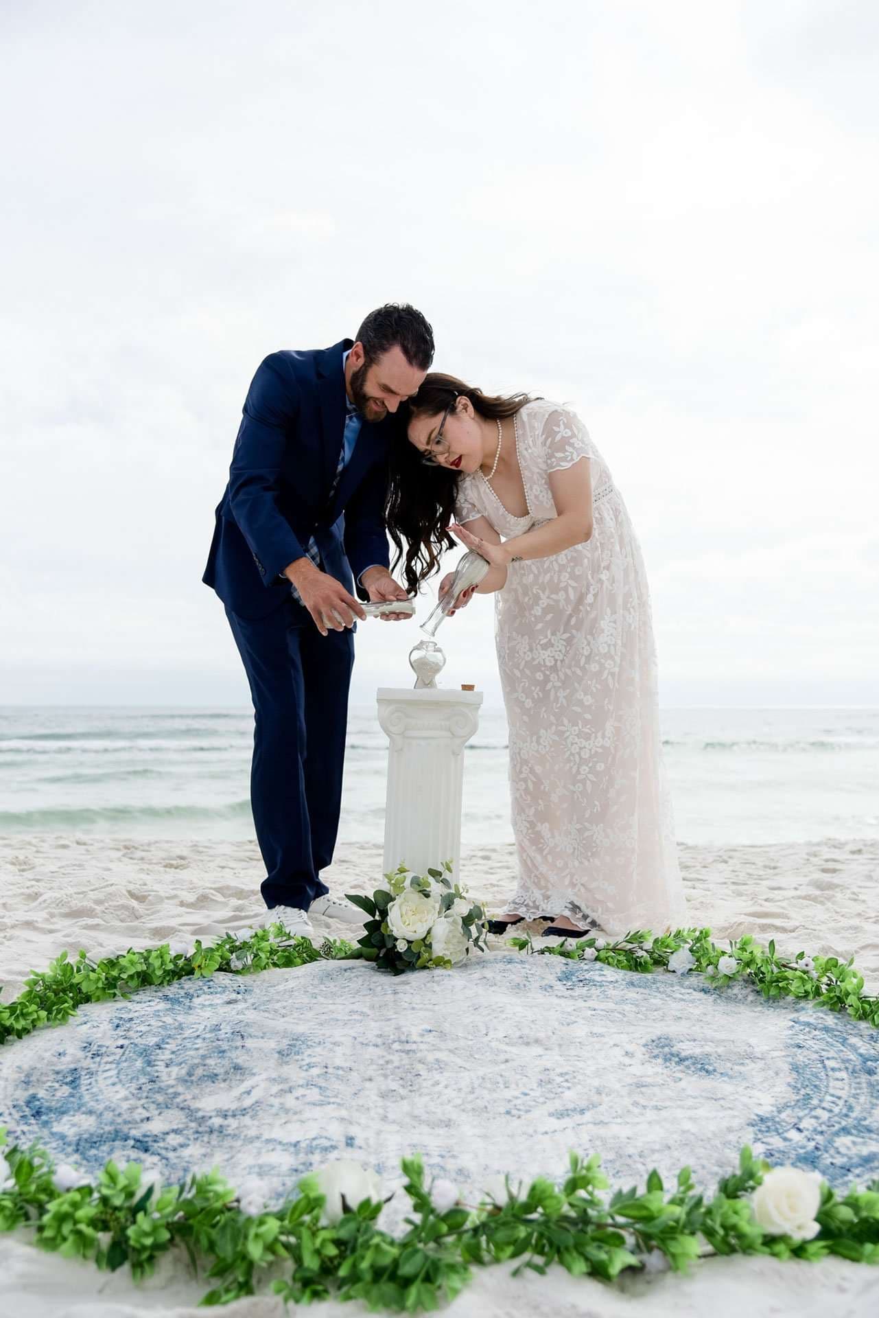 Couple pouring sand into a glass vessel during a unity ceremony on the beach, ringed by greenery and white roses