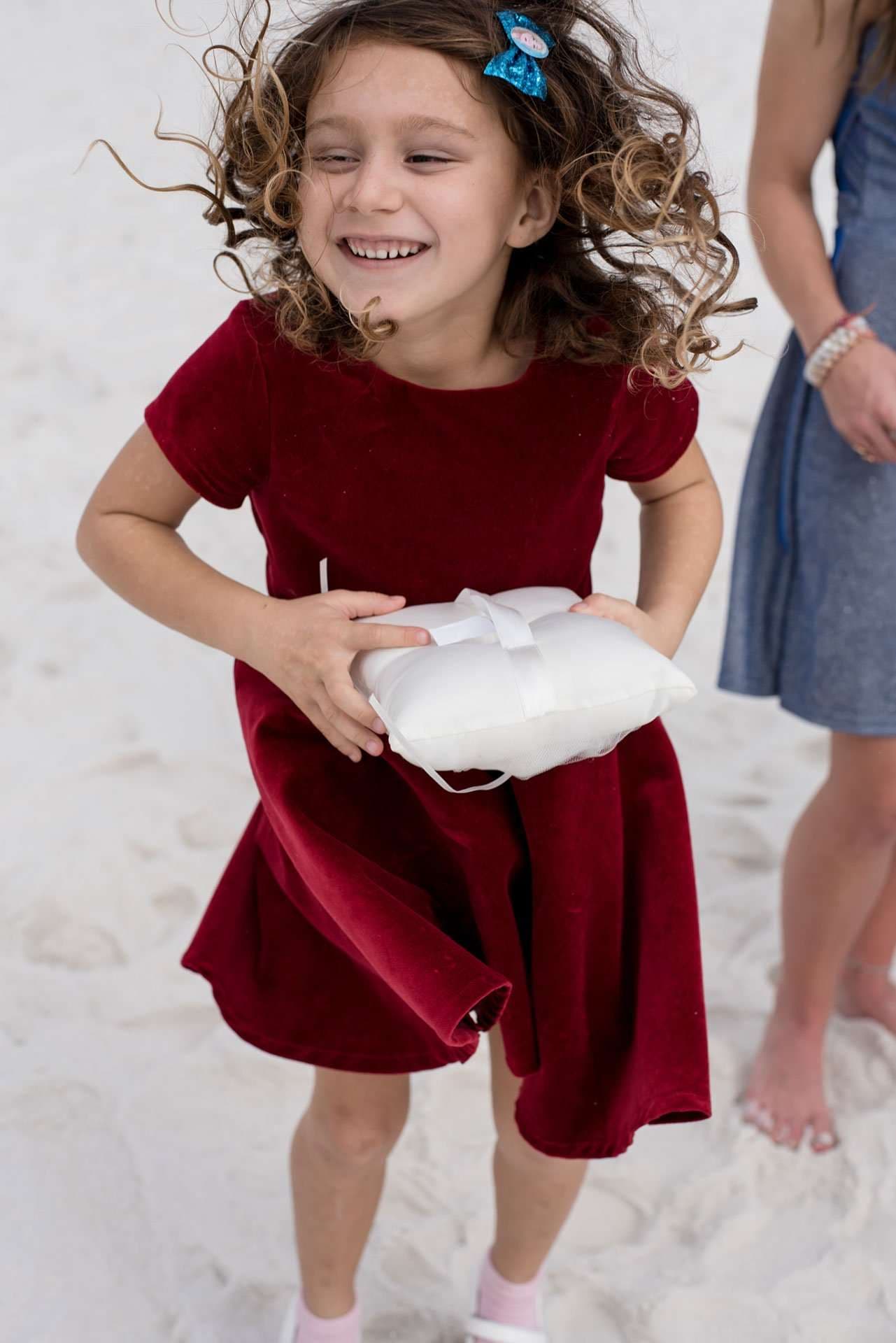 Laughing girl in a red velvet dress holding a white ring bearer pillow on the sand, her curly hair flying