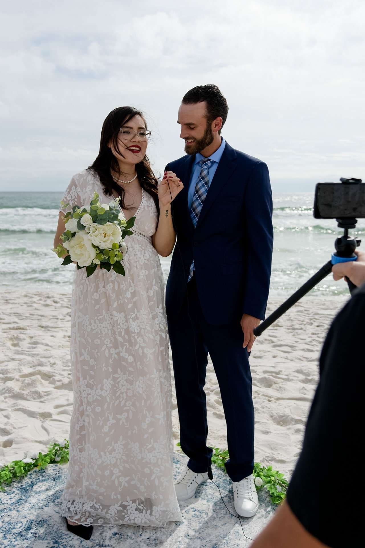 Couple exchanging rings on the beach, bride in a white lace gown holding a white bouquet, groom in a navy suit