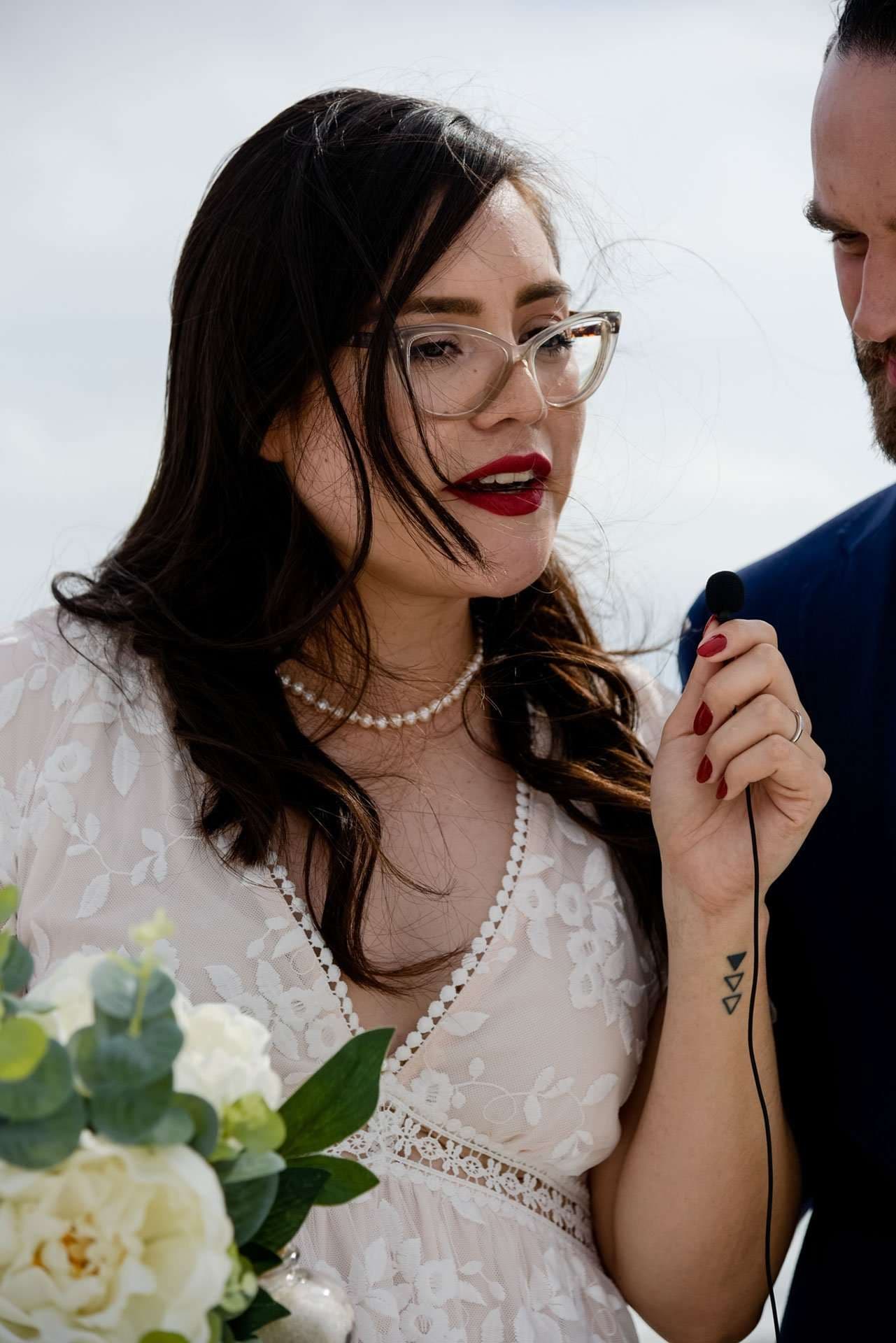 Bride in white lace dress and glasses speaks into a small lapel microphone, holding a white rose bouquet on Okaloosa Island
