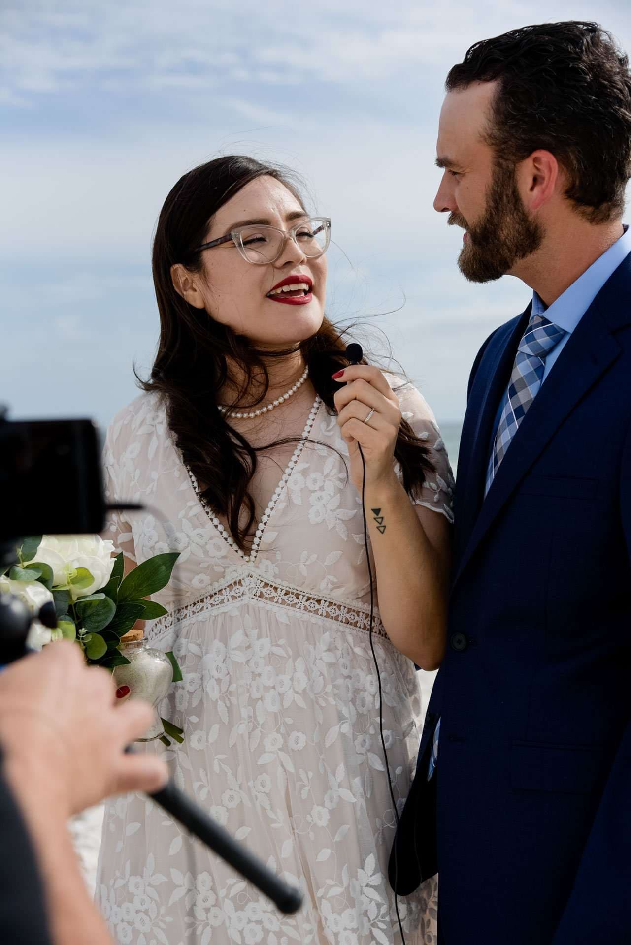 Bride in lace dress speaking into a small microphone while smiling groom in navy suit looks on, Okaloosa Island elopement