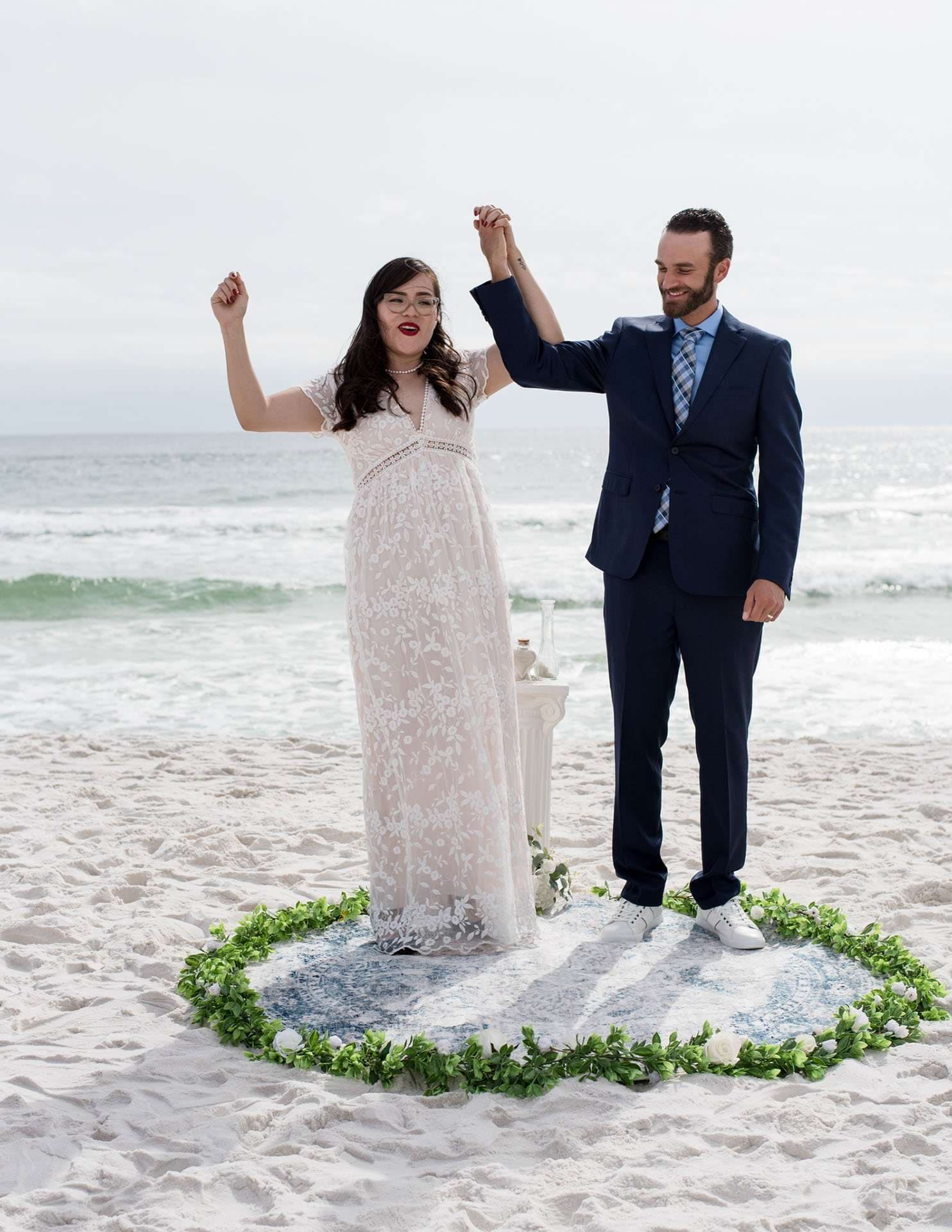 Couple raising joined hands in celebration on a beach inside a circle of greenery, bride in a white lace gown