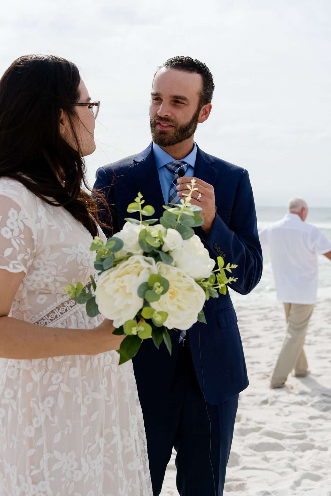 Bearded man in a navy suit talks with a woman in a white lace dress holding a white peony bouquet on Okaloosa Island beach