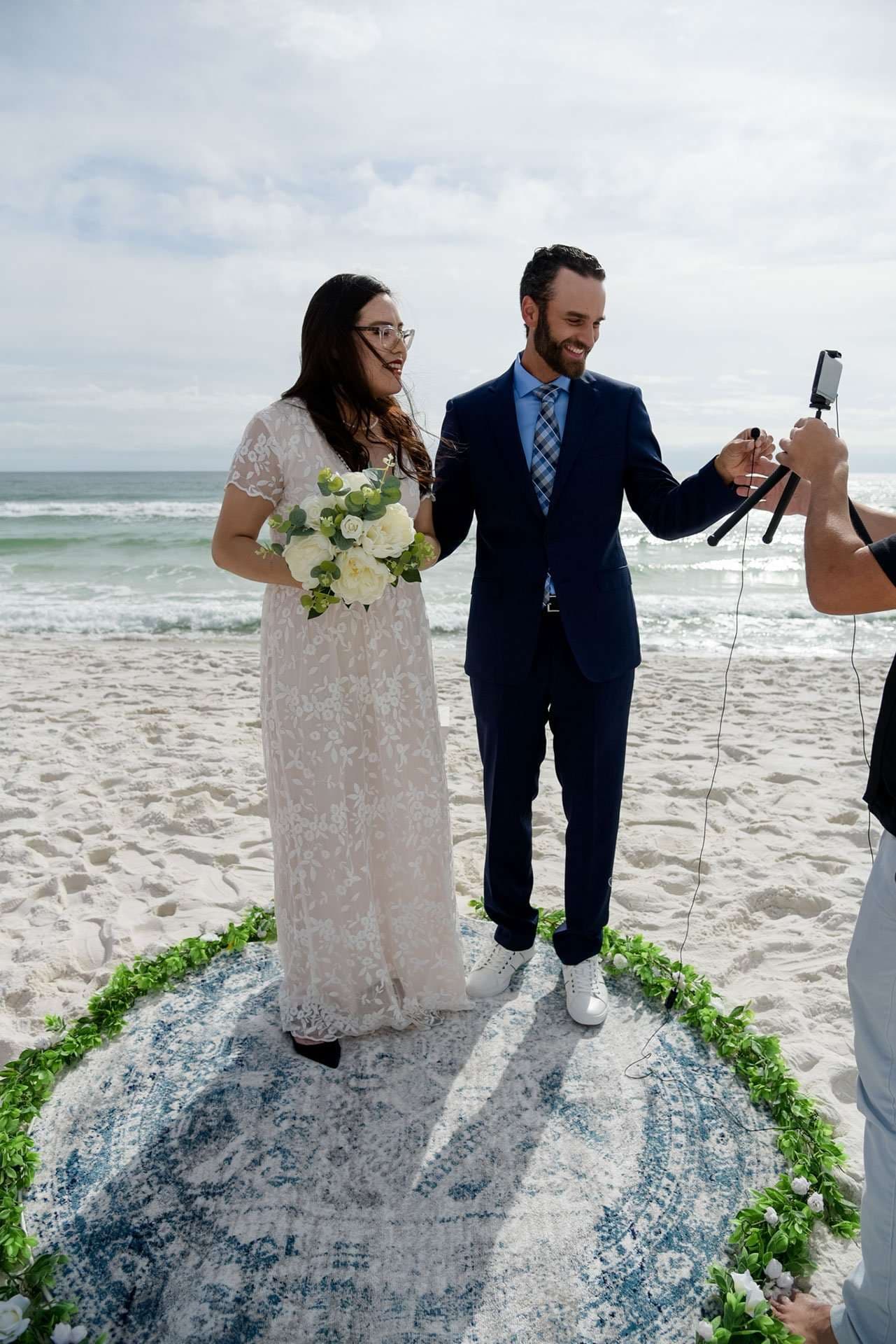 Couple in a lace gown and navy suit stand on a round rug at the beach while someone films them