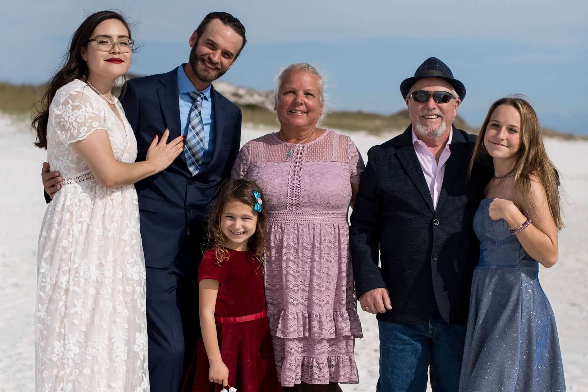Family of six in formal attire posing together on a white sand beach at Okaloosa Island, smiling at the camera