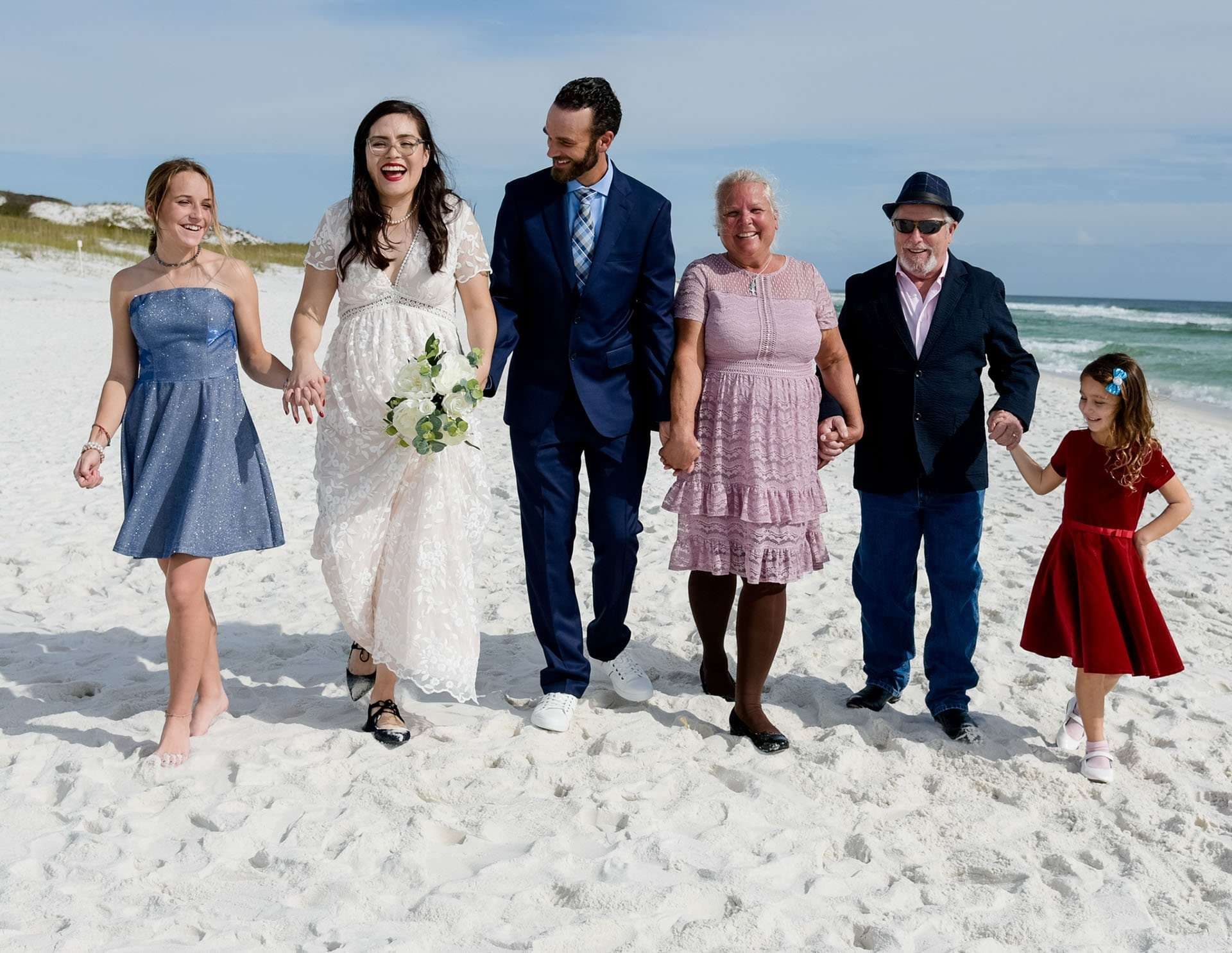 Bride and groom walk hand in hand along a white sand beach with four smiling family members, Okaloosa Island