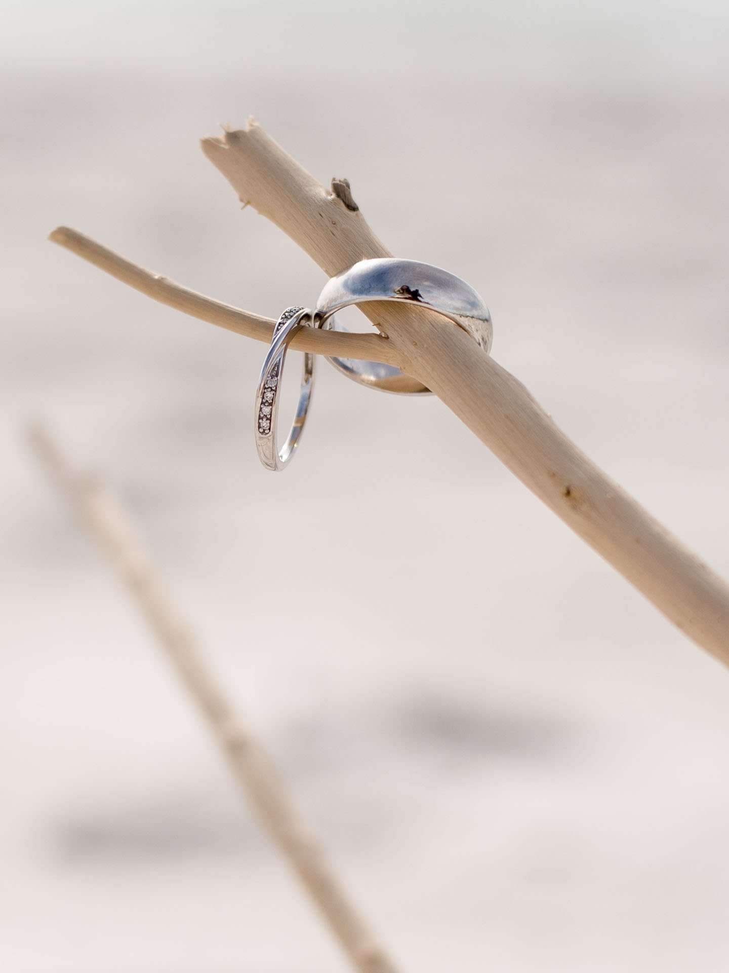 Two silver wedding bands resting on a driftwood branch against a soft, blurred sandy beach background