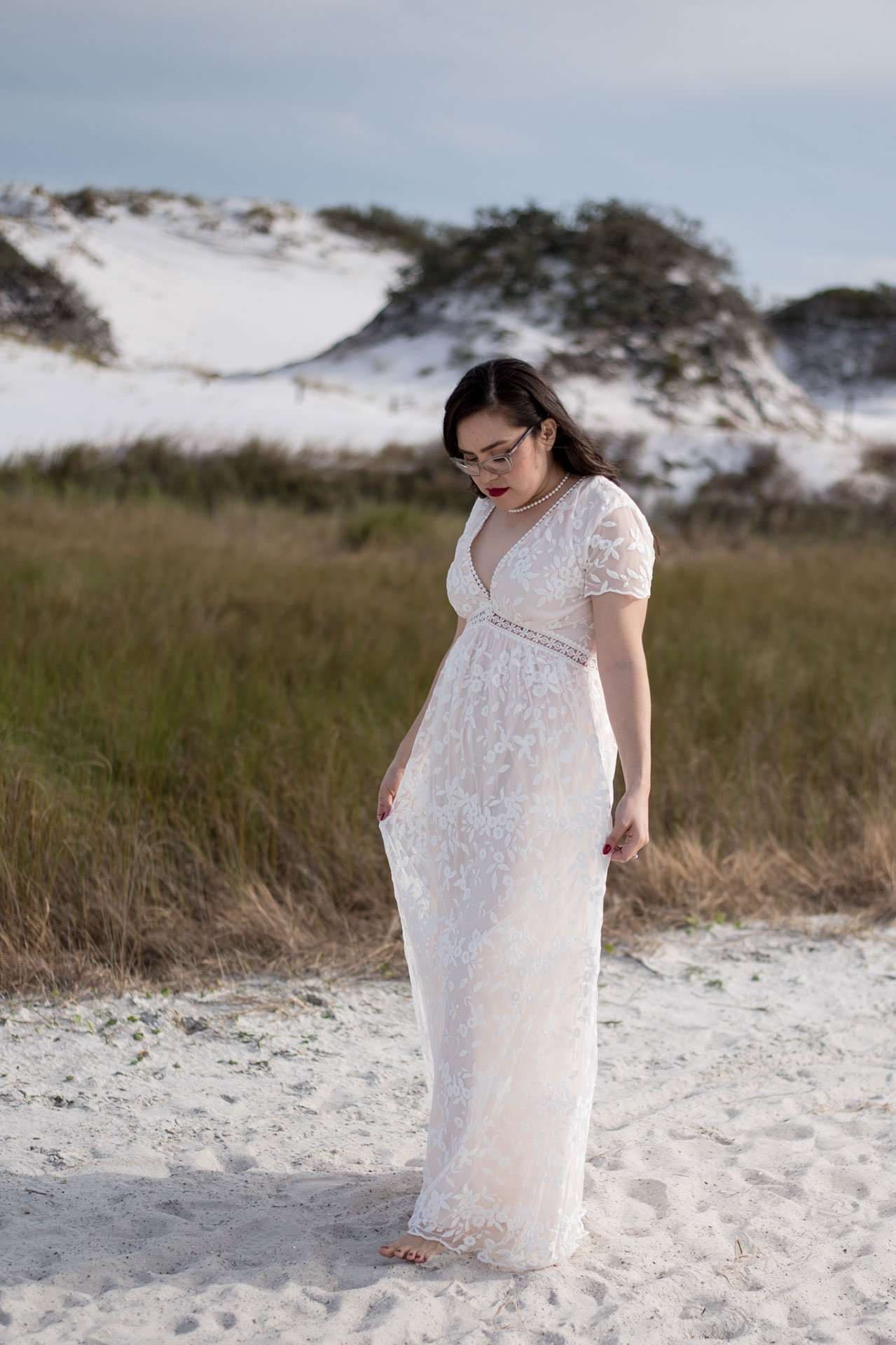 Barefoot woman in a white lace gown and glasses looking down on white sand with grassy dunes behind her