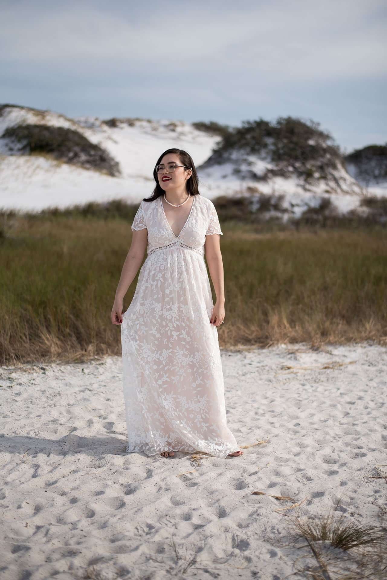 Woman in a white lace gown and pearl necklace stands on white sand dunes with grasses behind her