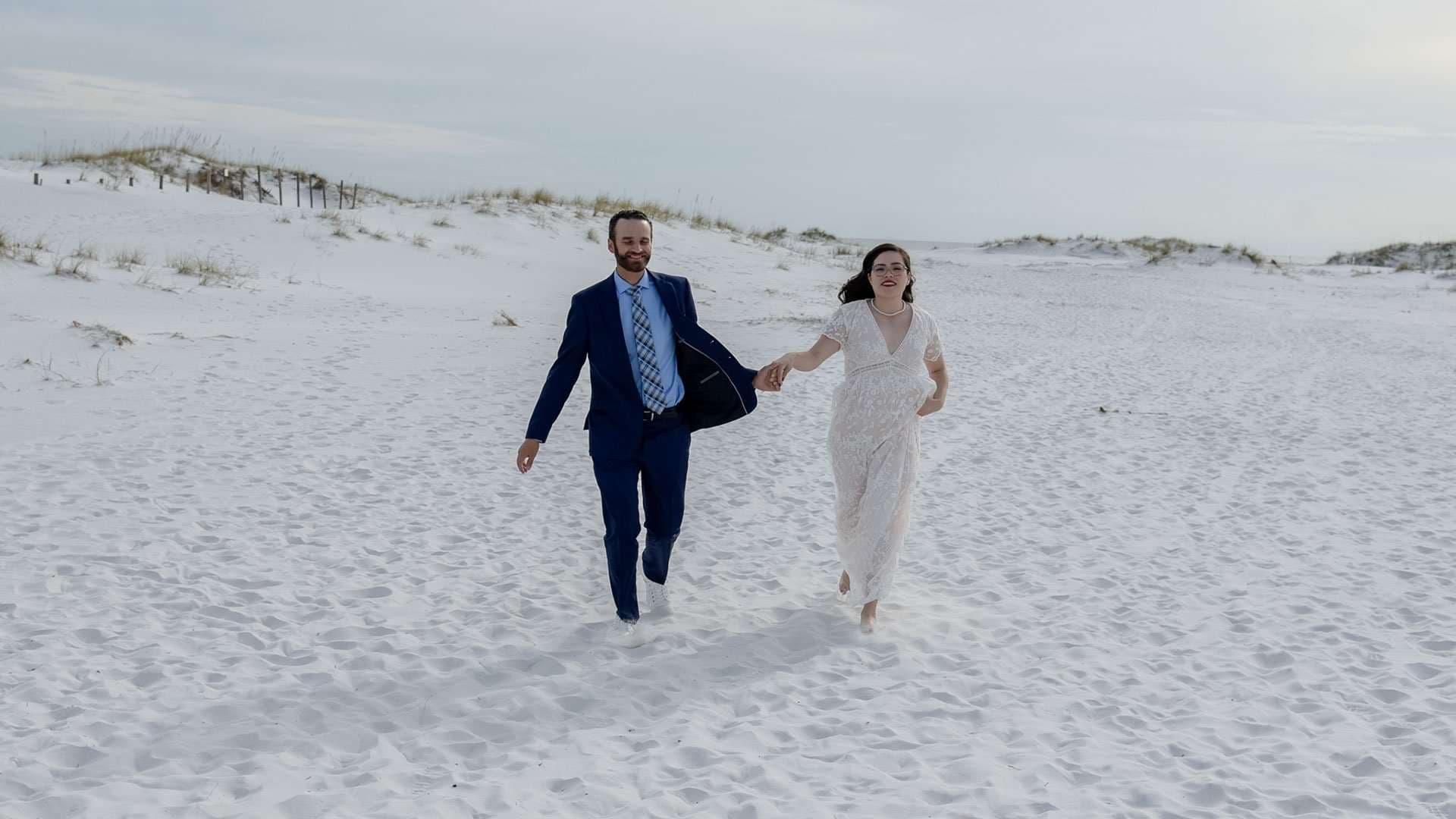 Couple holding hands and running barefoot across white sand dunes on Okaloosa Island, he in a navy suit, she in white lace