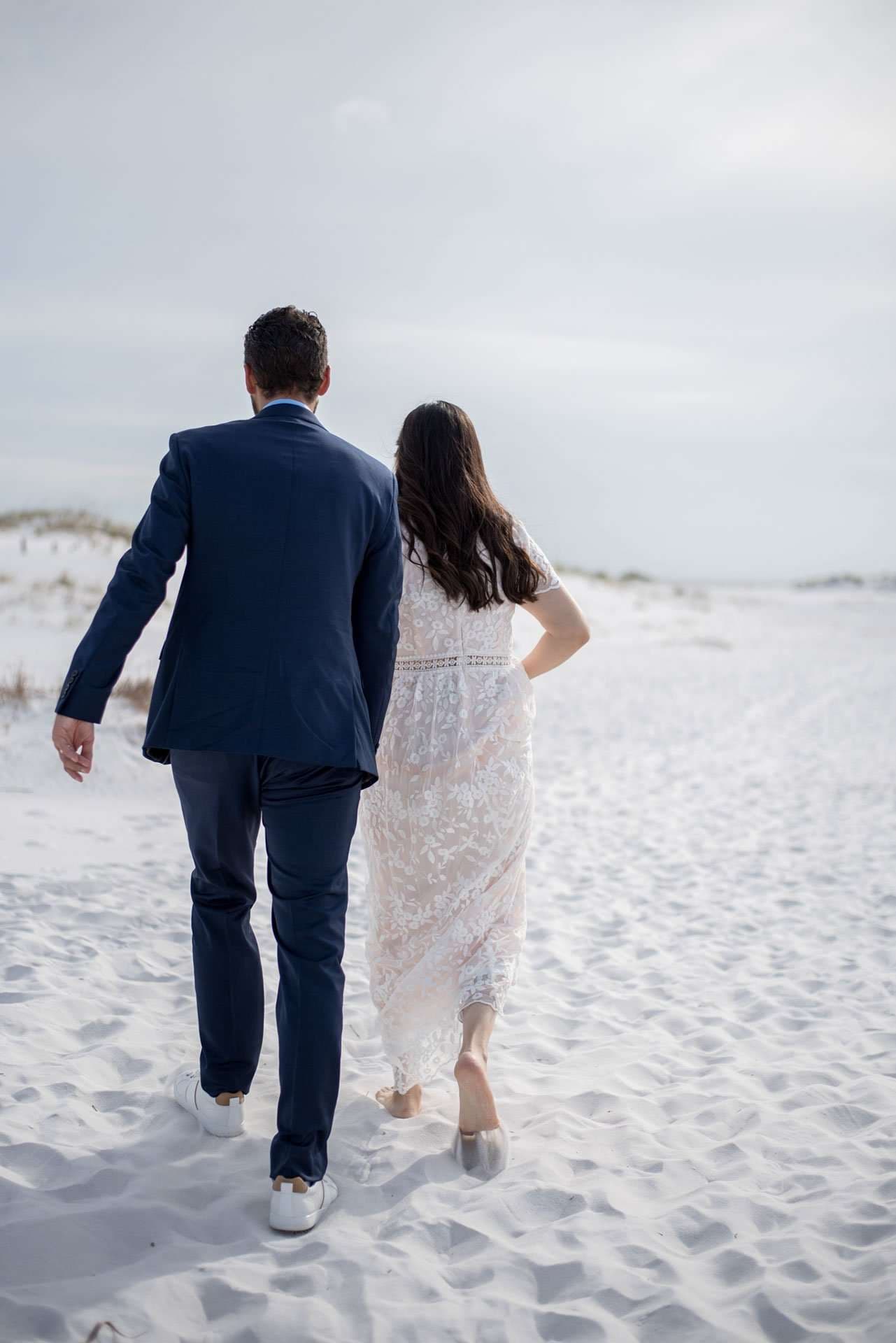 Couple in a navy suit and white lace dress walking barefoot away across white sand dunes on Okaloosa Island