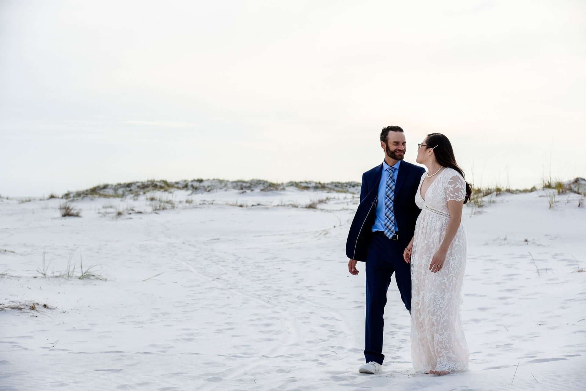 Couple in a navy suit and white lace gown walking and smiling at each other on white sand dunes on Okaloosa Island