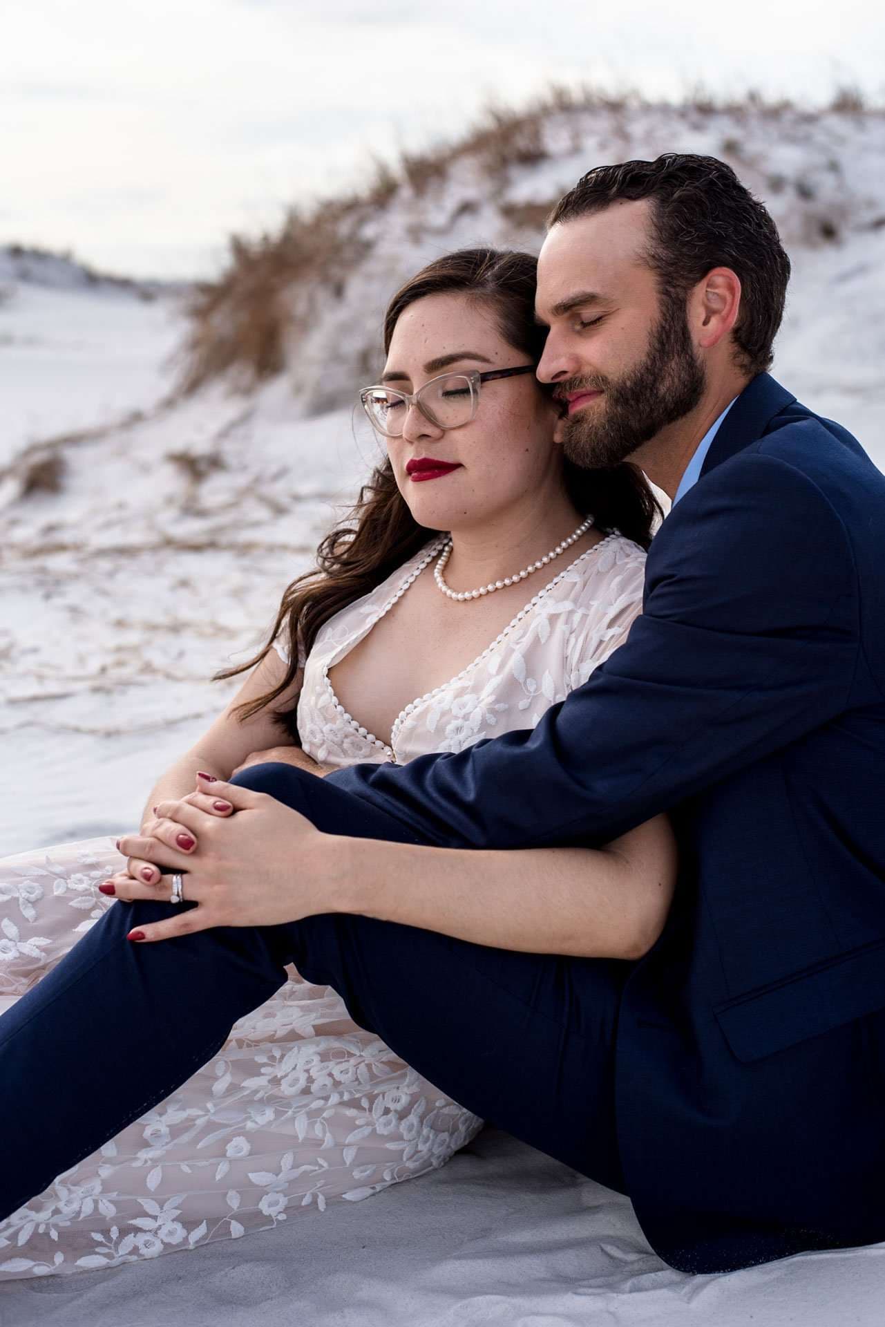Couple seated on white sand dunes, man in navy suit embracing woman in lace gown and pearl necklace from behind