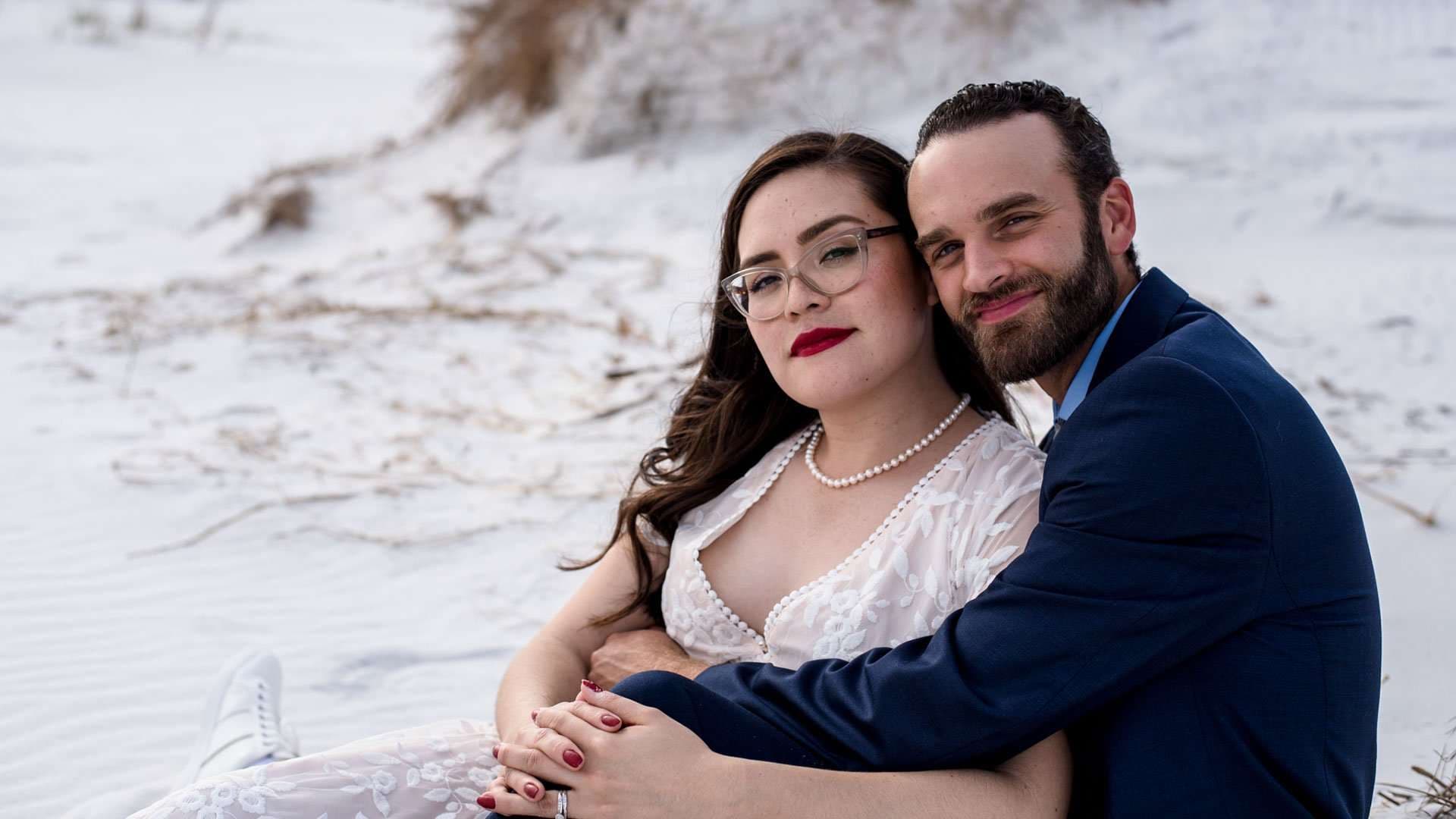Couple in wedding attire embracing on white sand at Okaloosa Island, she wears glasses and pearls, he a navy suit