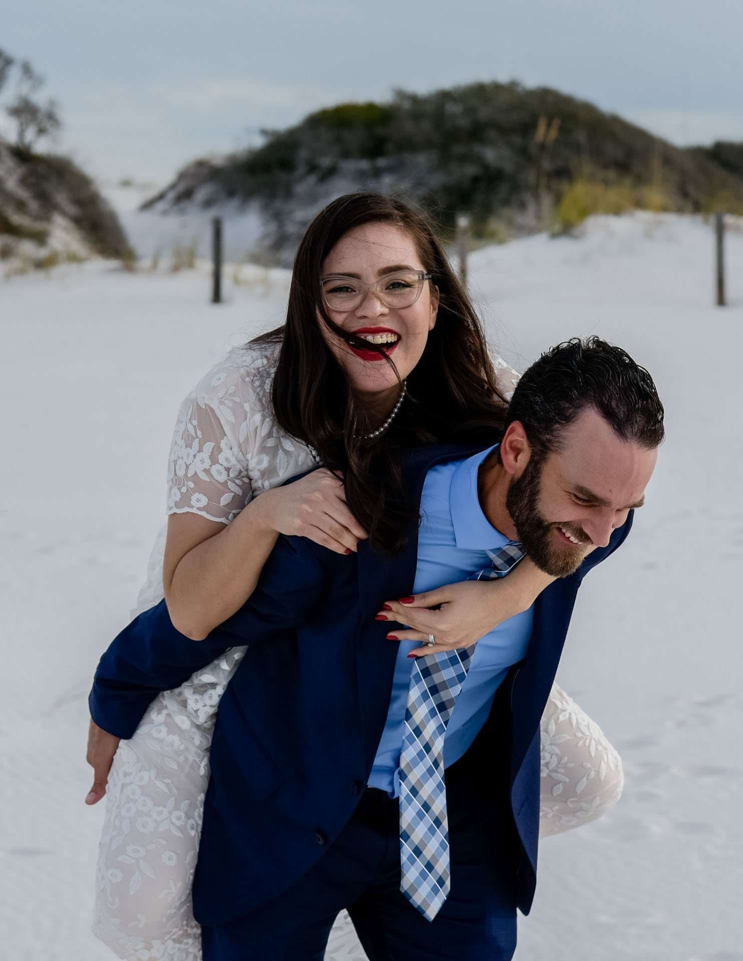 Laughing bride in a white lace dress riding piggyback on her smiling groom in a blue suit on white sand dunes