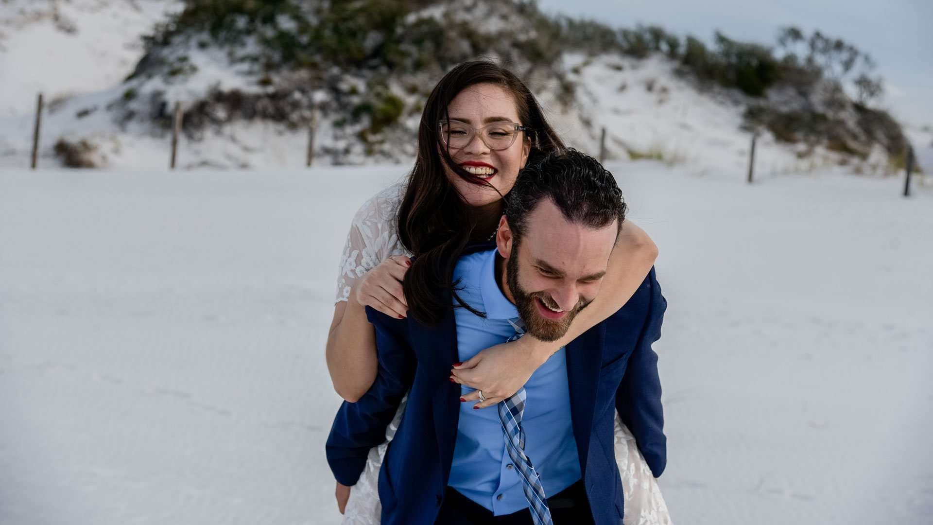 Laughing bride in a white lace dress rides piggyback on her groom in a navy jacket across white sand dunes