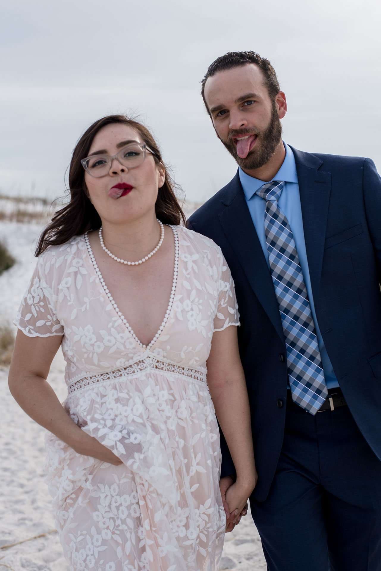 Couple sticking out their tongues playfully on a white sand beach, she in a lace dress, he in a navy suit
