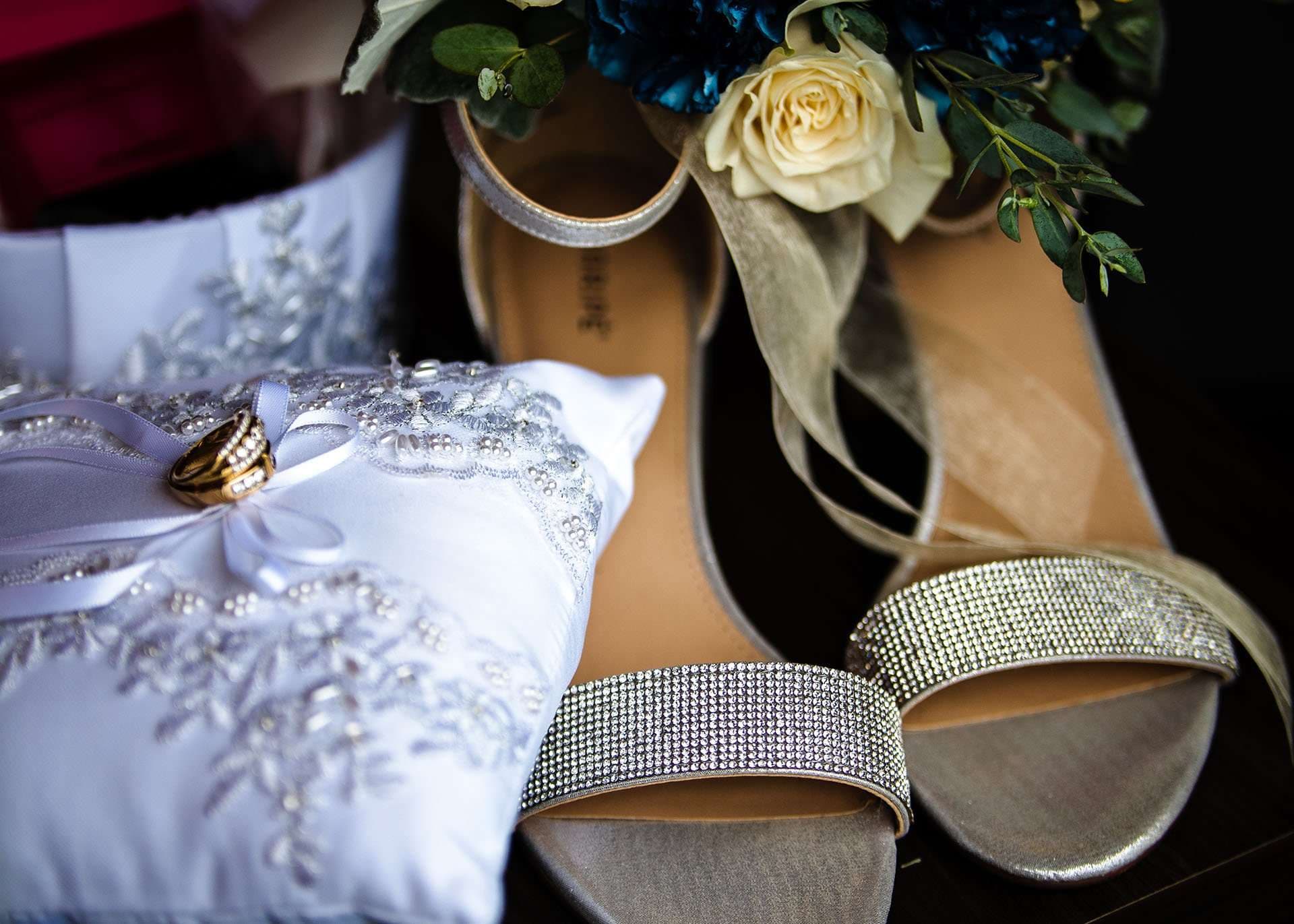 Gold wedding ring on a white beaded ring pillow beside silver rhinestone heels and a cream rose bouquet