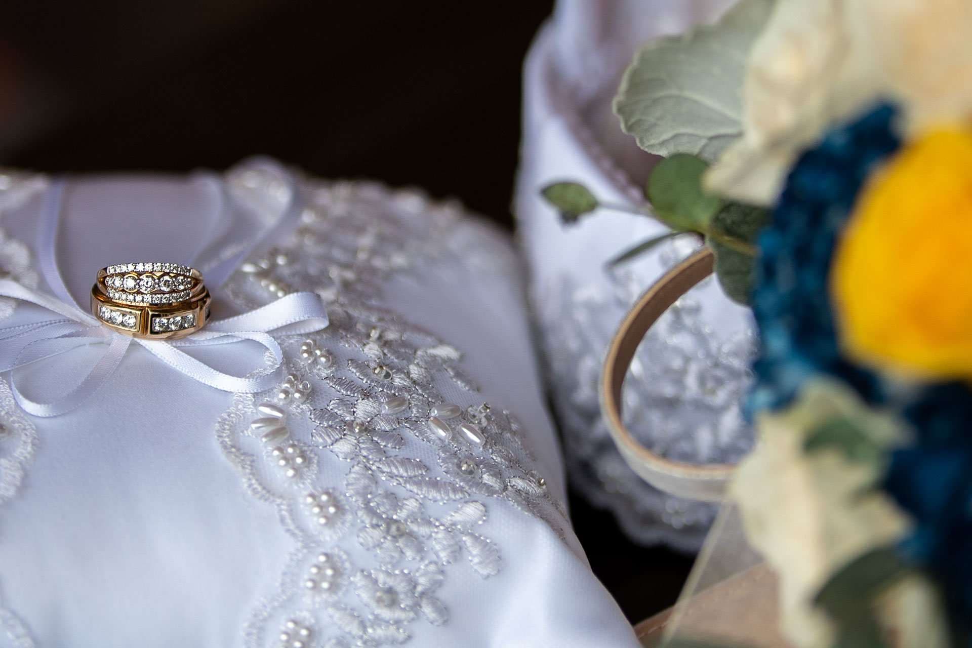 Gold diamond wedding rings tied with a white ribbon to a beaded lace pillow beside a blue and yellow bouquet