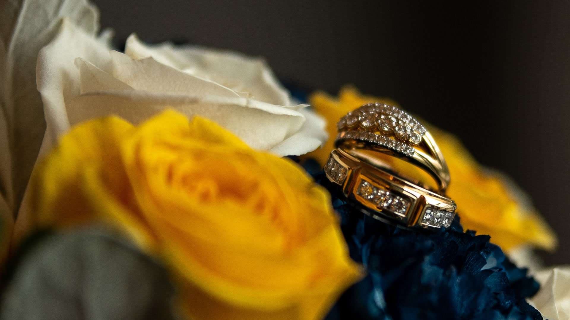 Two gold diamond wedding rings resting on a bouquet of yellow and white roses with dark blue flowers