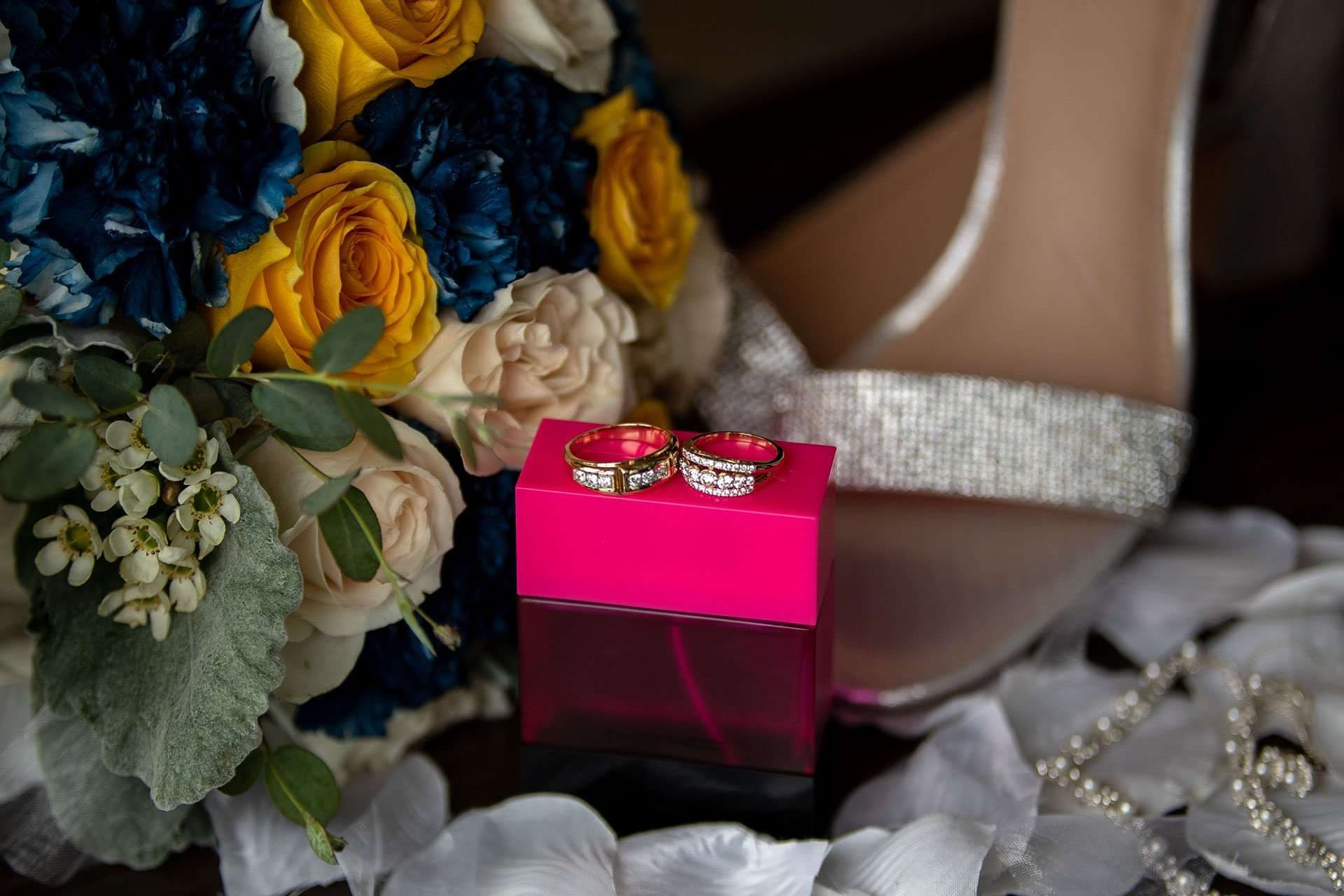 Two gold wedding bands resting on a hot pink box beside a navy and yellow bouquet and a sparkly silver sandal