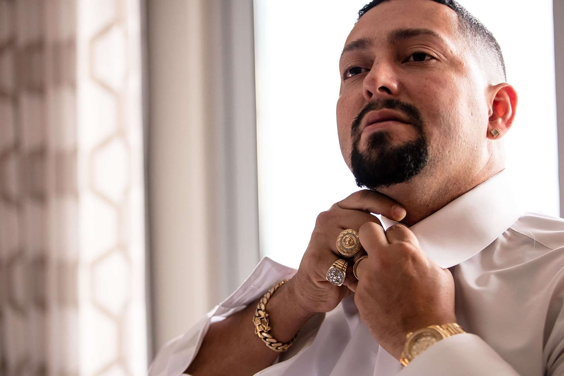 Groom in a white dress shirt adjusting his collar, wearing gold rings, a bracelet and a gold watch by a window