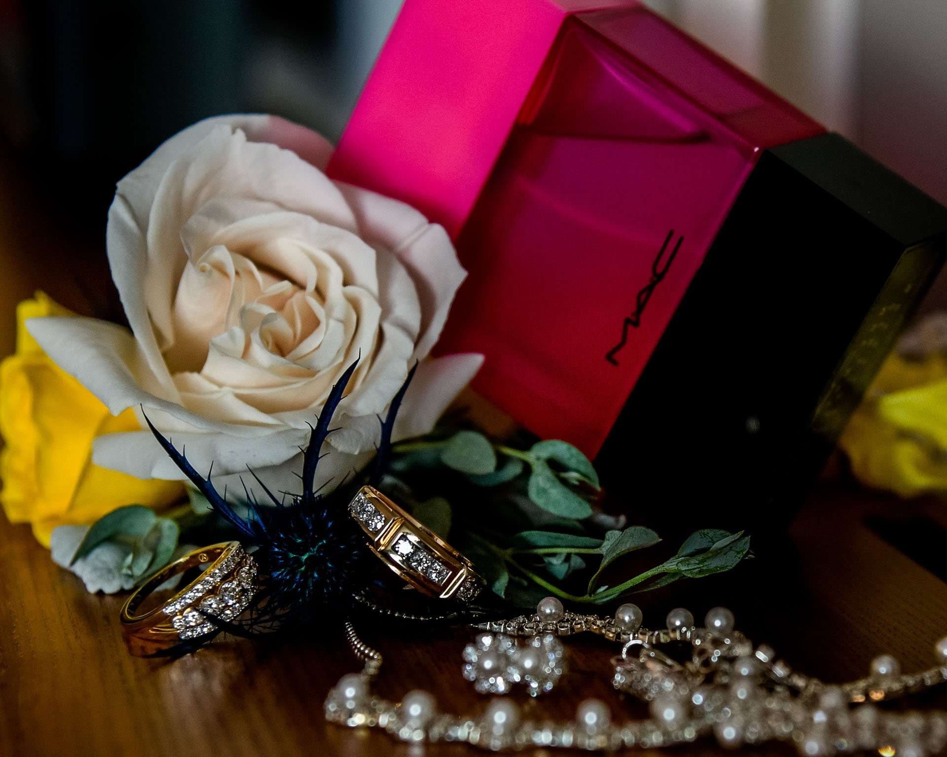 Two diamond wedding bands and a pearl necklace beside a cream rose and a pink MAC box on a wooden table