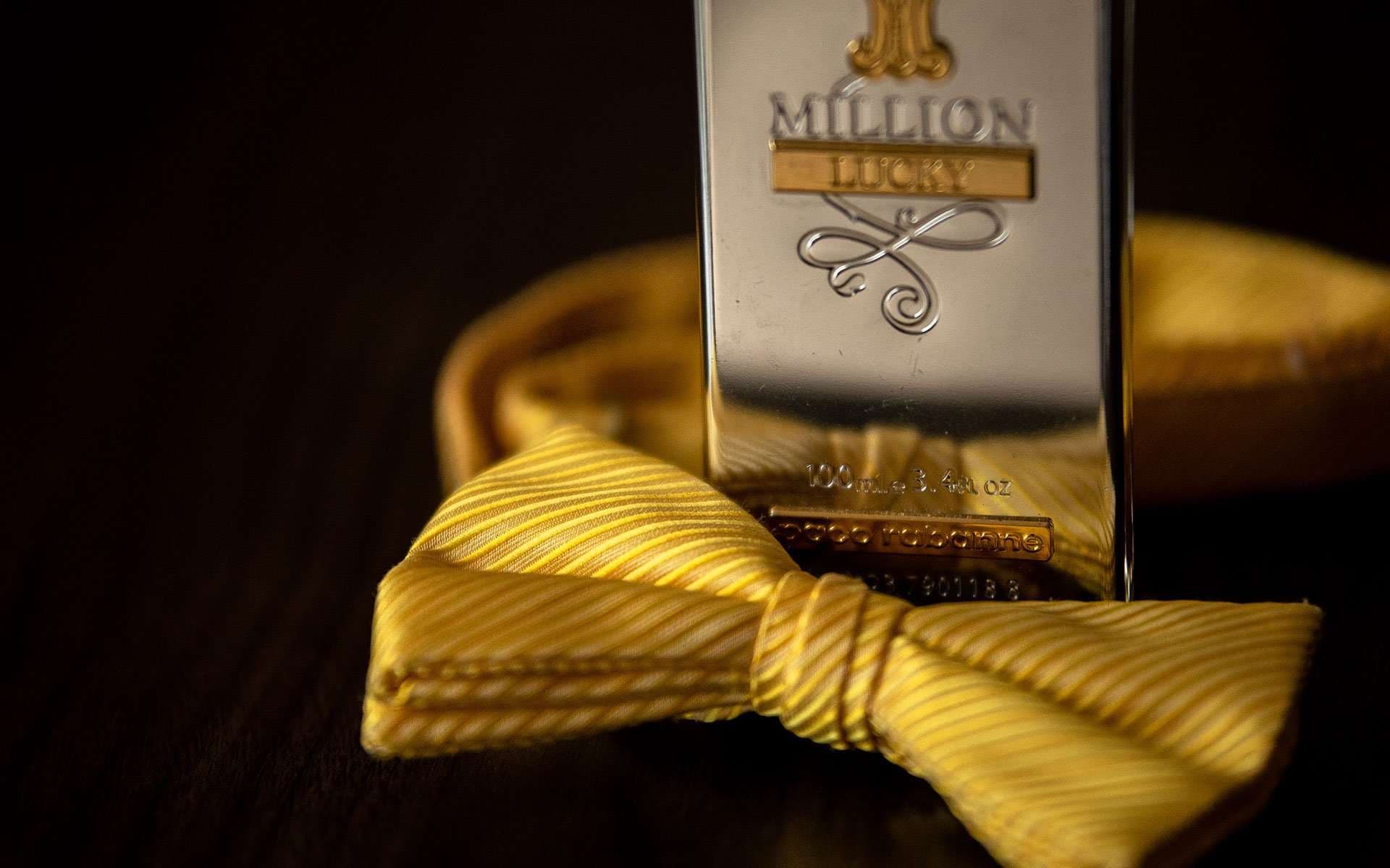 Yellow striped silk bow tie resting beside a metallic 1 Million Lucky cologne bottle on a dark wooden surface