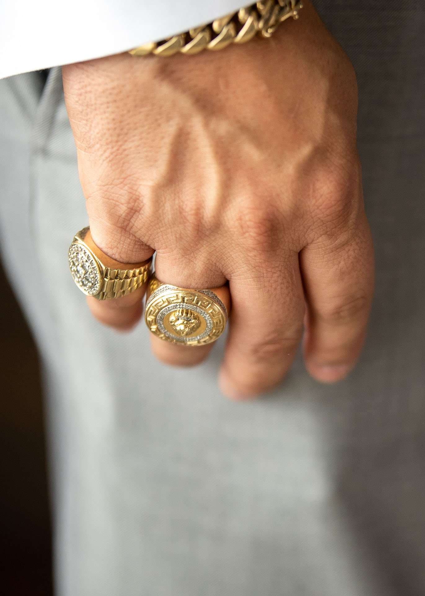 Close-up of a hand in a gray suit wearing two gold signet rings and a gold chain bracelet