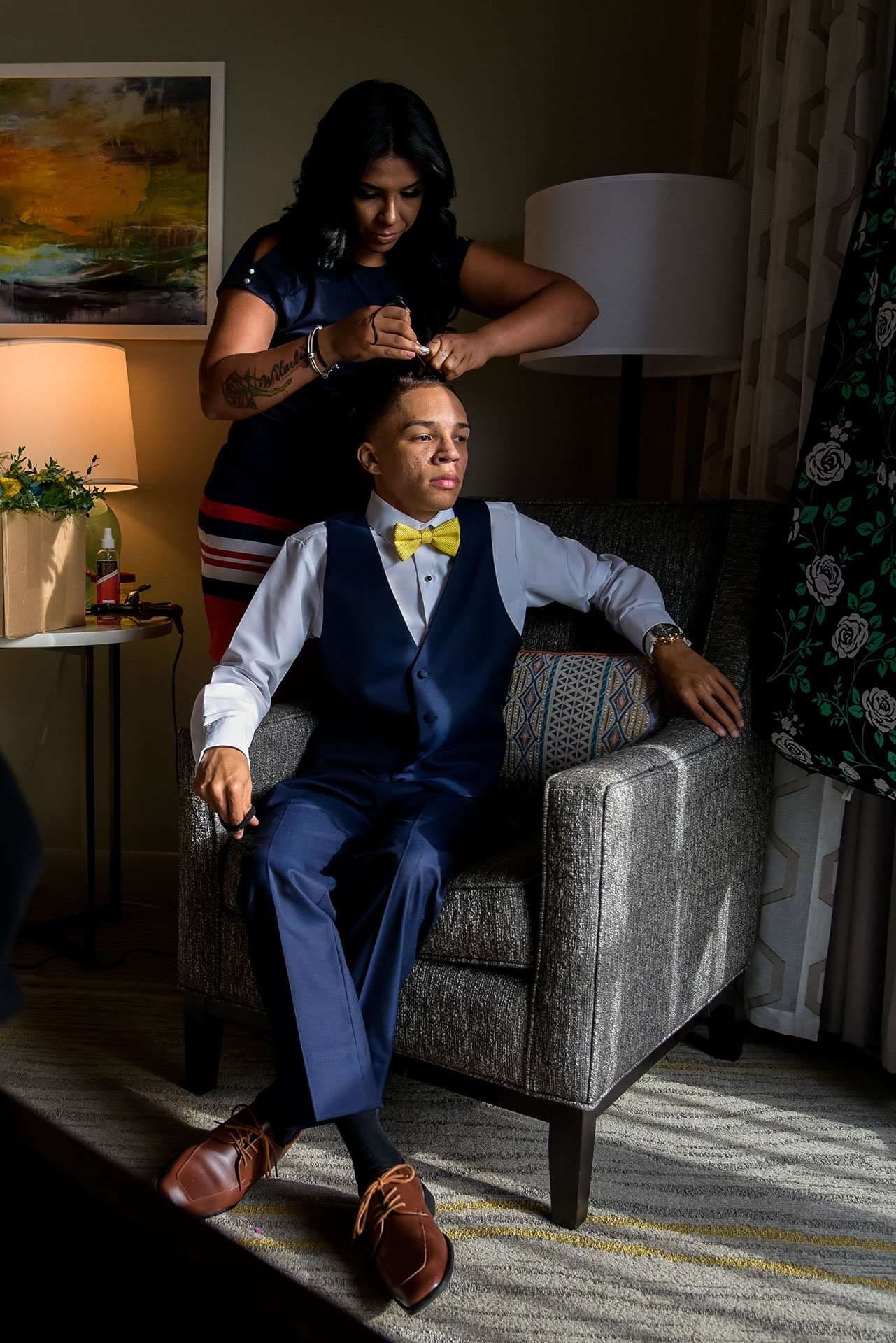 Woman adjusting a seated young man's hair as he sits in a armchair wearing a navy vest, yellow bow tie and brown shoes