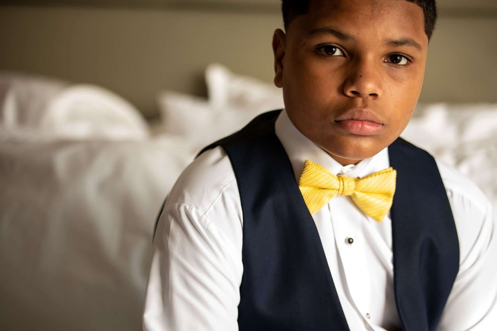 Serious young man in a navy vest, white shirt and yellow bow tie sitting on a white-linen bed
