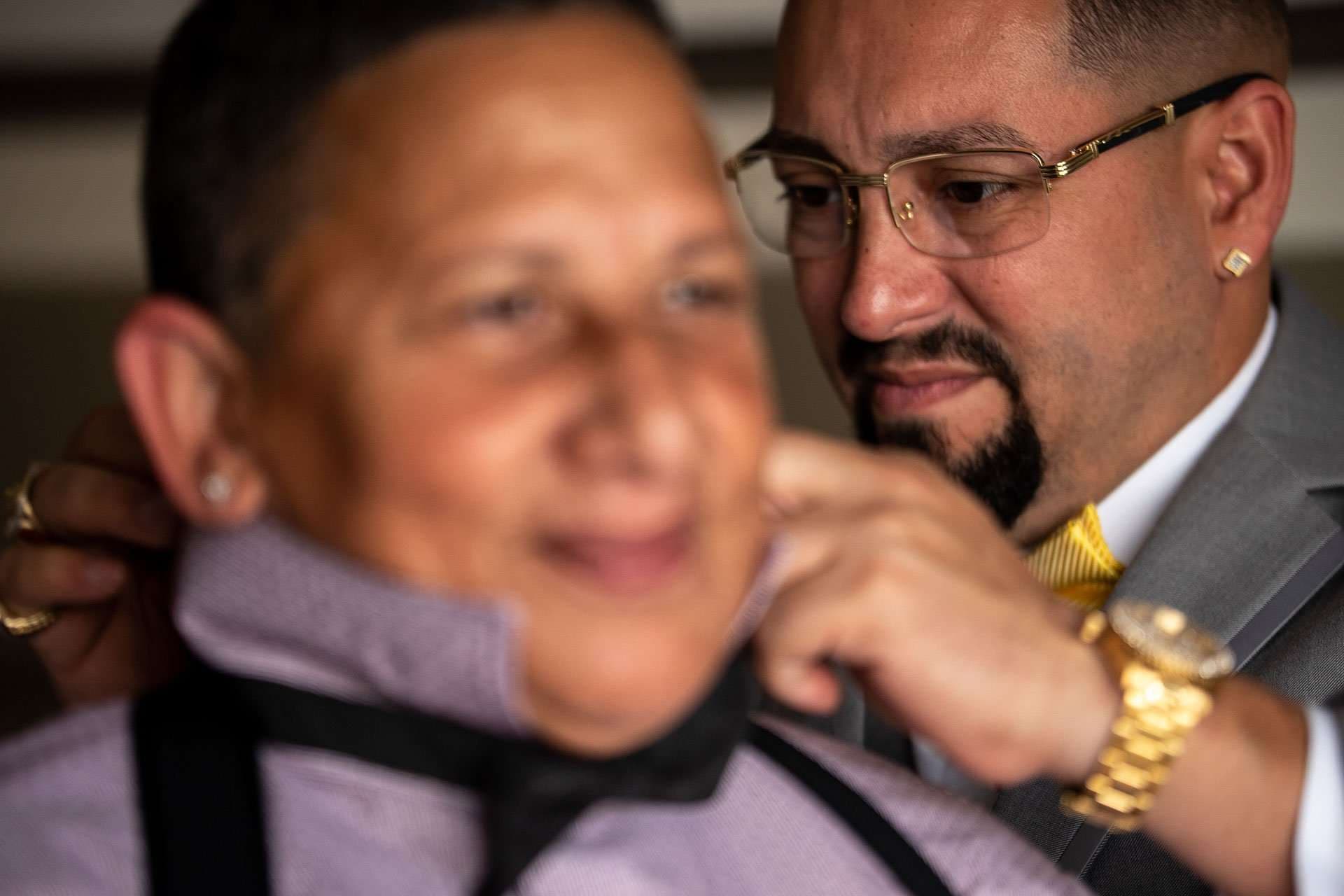 Man in glasses and gray suit adjusts a black bow tie on a smiling person in a lavender shirt and suspenders