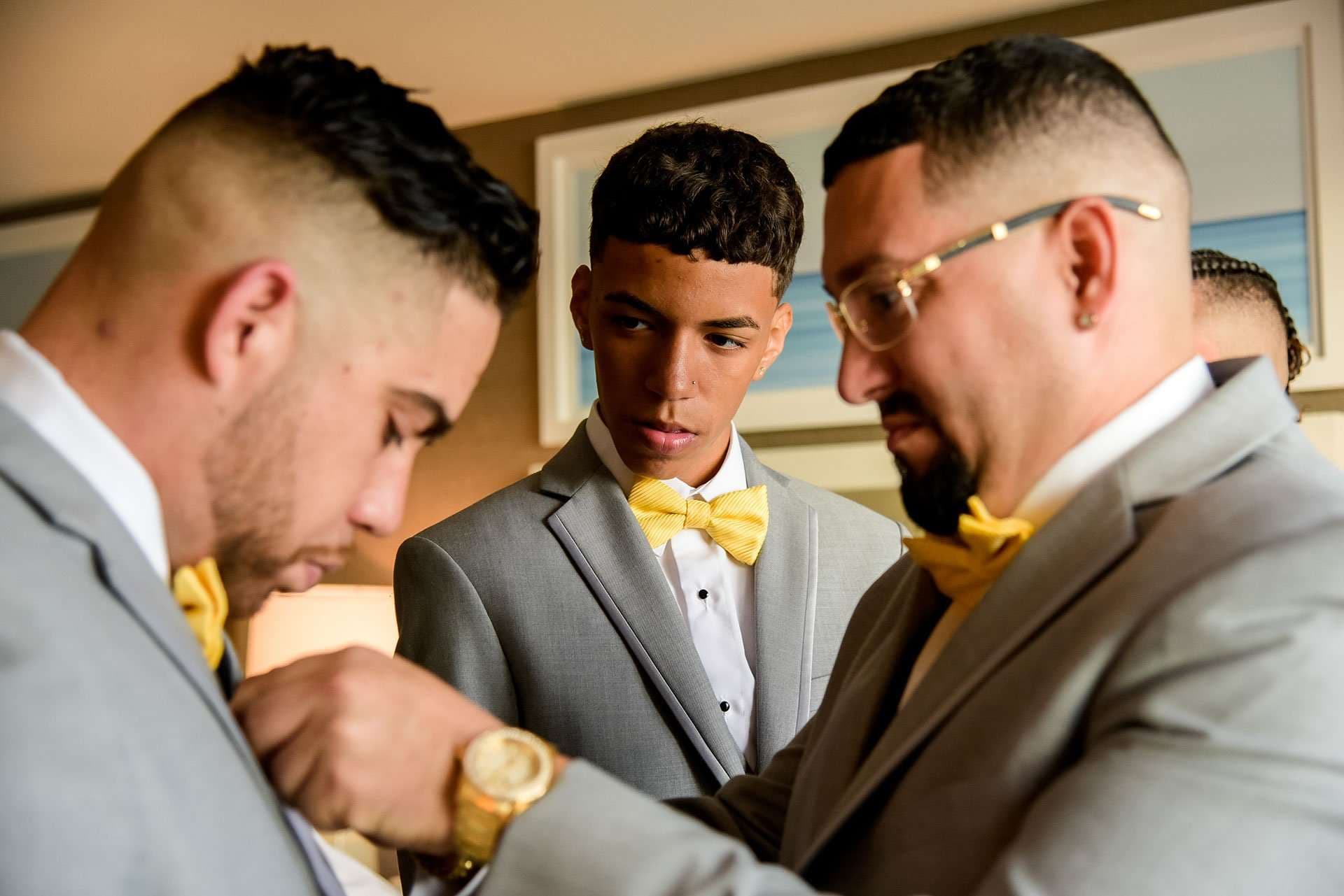 Groomsmen in gray suits and yellow bow ties getting ready, one adjusting another's lapel while a young man watches