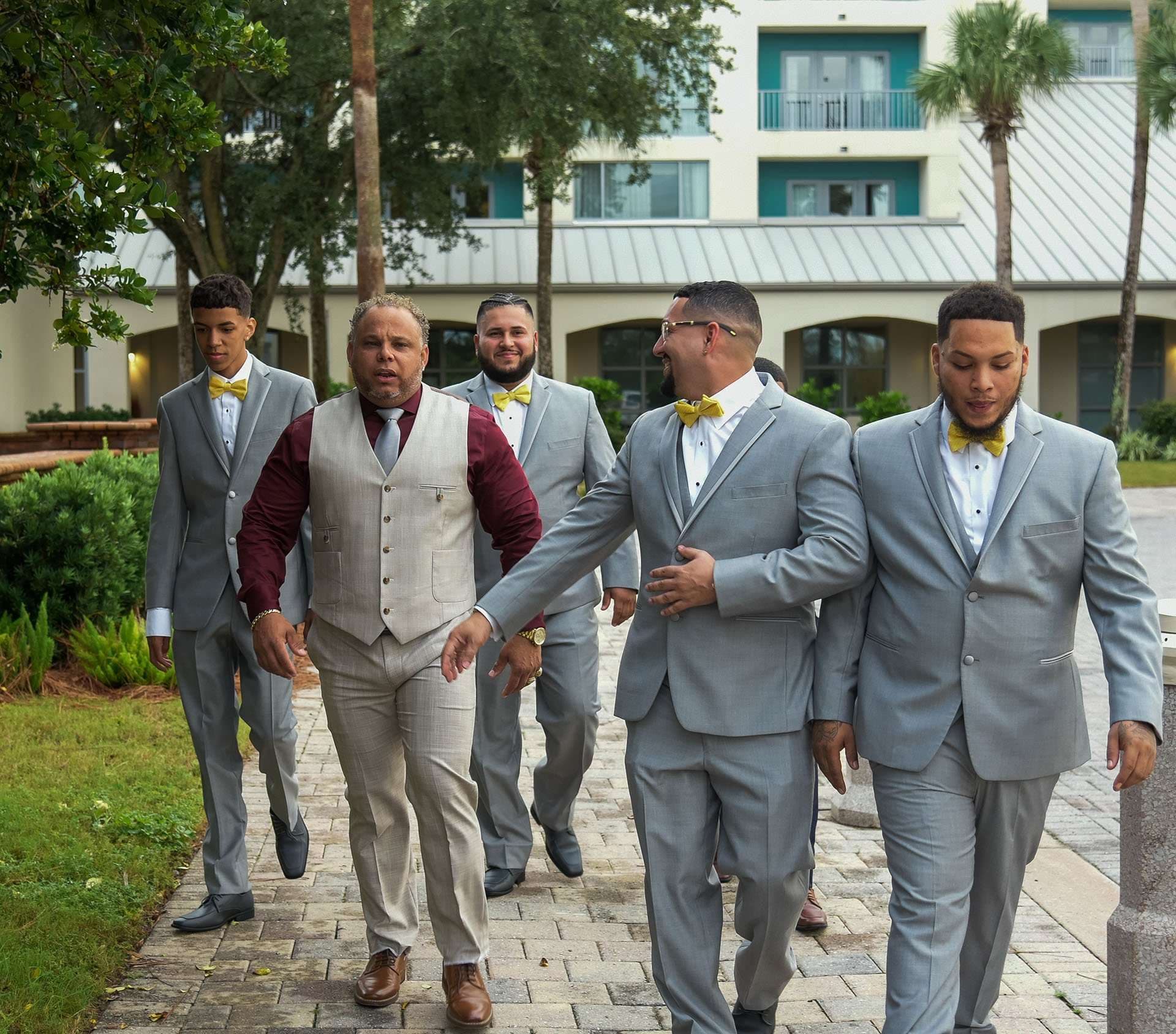 Five men in gray suits and yellow bow ties walking together along a brick path outside a resort