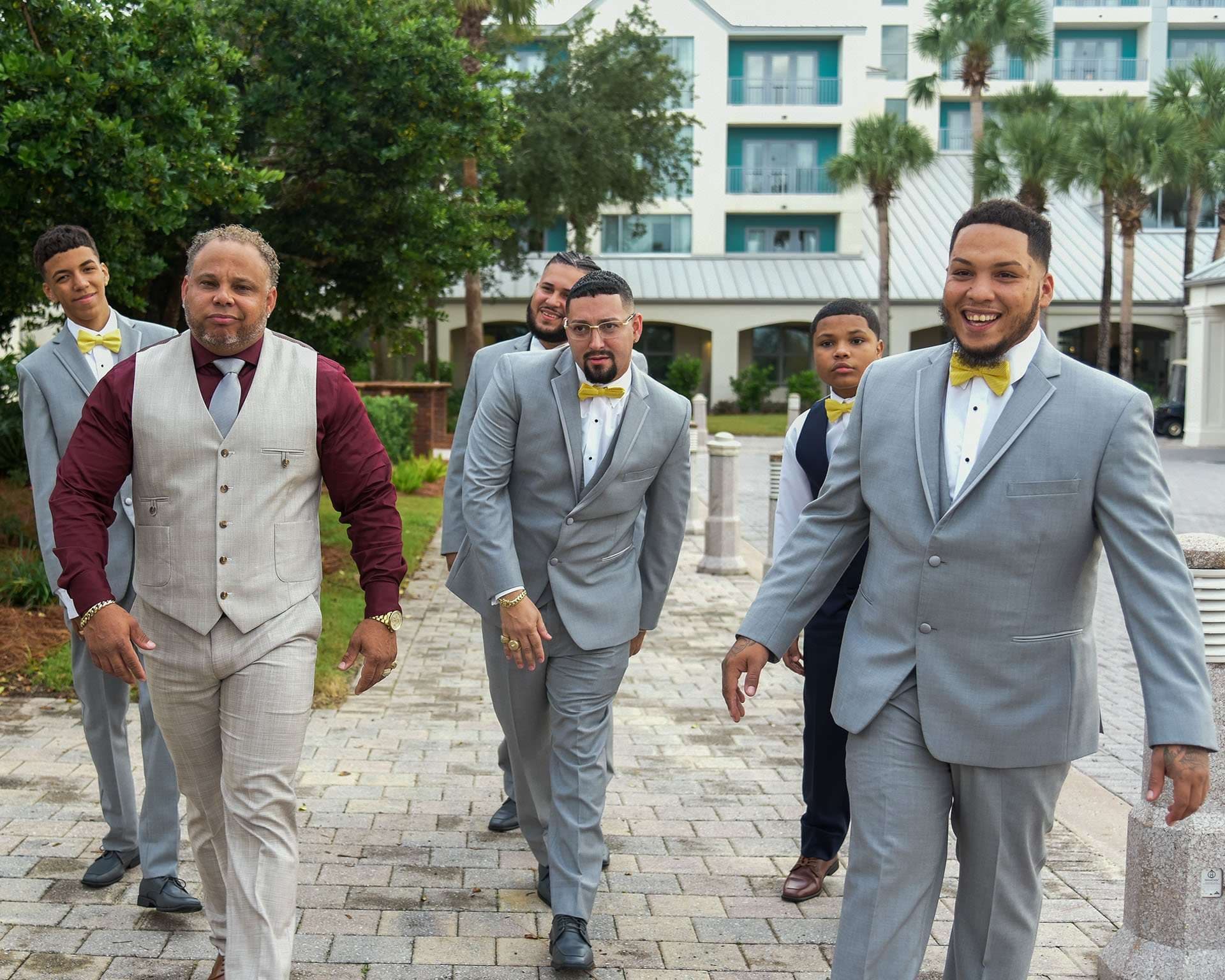 Smiling groomsmen in gray suits and yellow bow ties walking along a brick path outside the resort