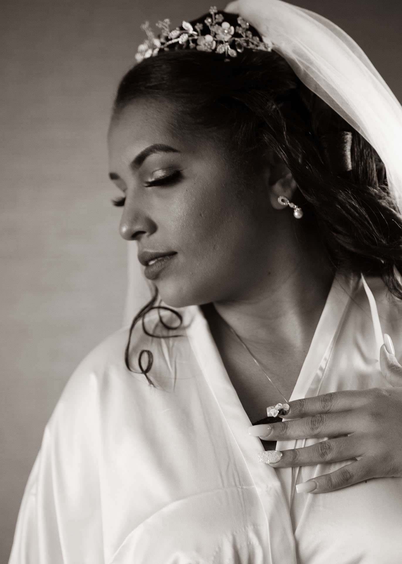 Black-and-white portrait of a bride in a satin robe, veil and floral tiara, eyes closed, hand at her collar
