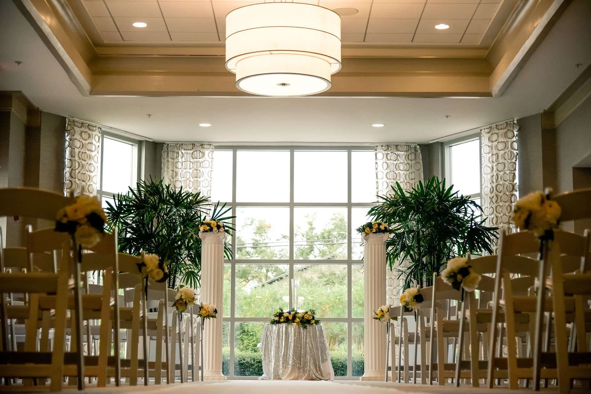 Indoor wedding ceremony aisle with white chairs, yellow floral bouquets, white columns and palms before a large bright window
