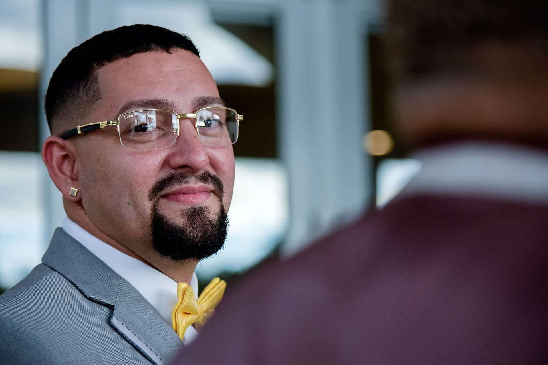 Smiling man in gold-rimmed glasses, gray suit and yellow bow tie glancing over his shoulder at a blurred guest