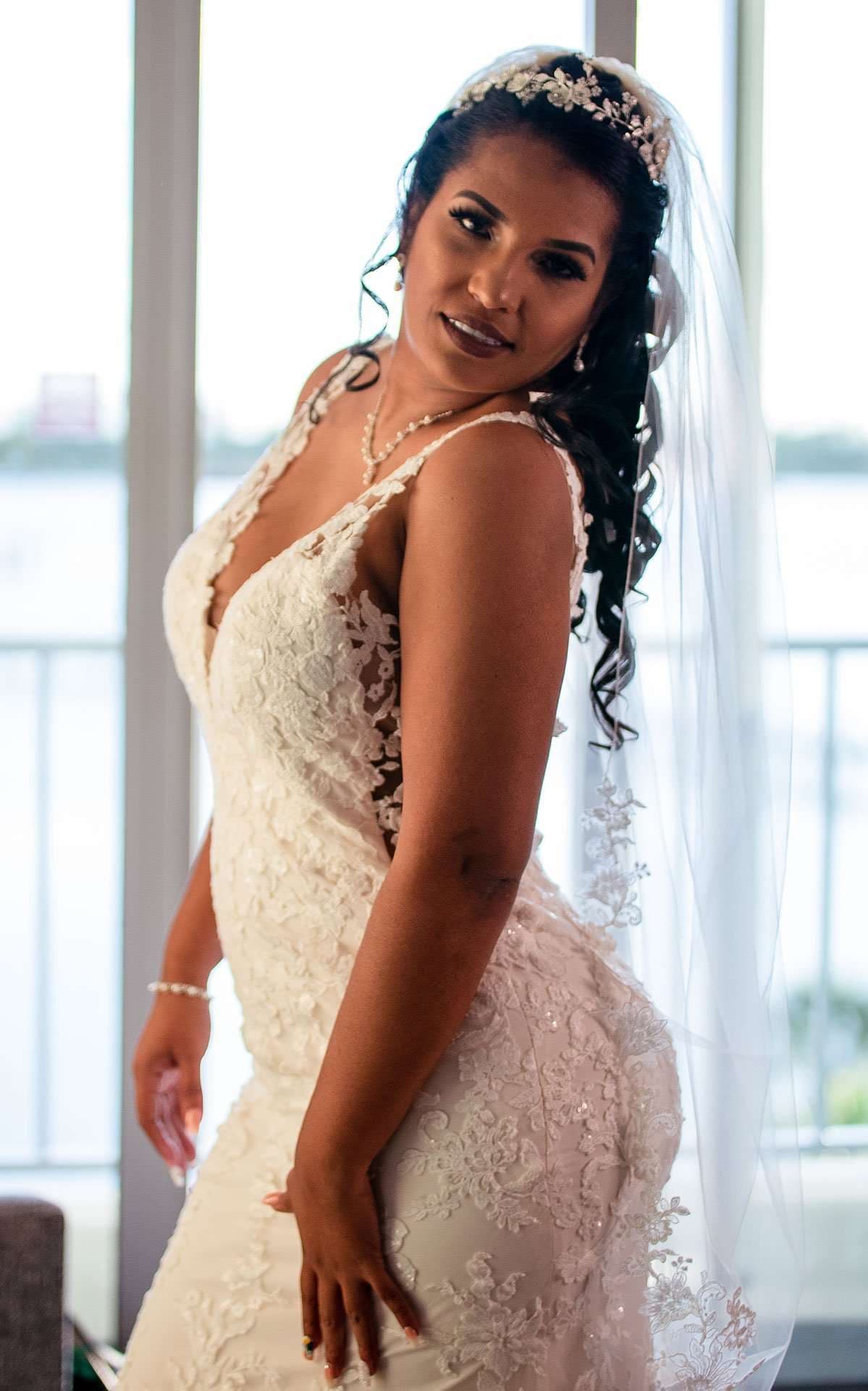 Bride in a white lace gown, floral tiara and long veil posing beside tall windows overlooking the water