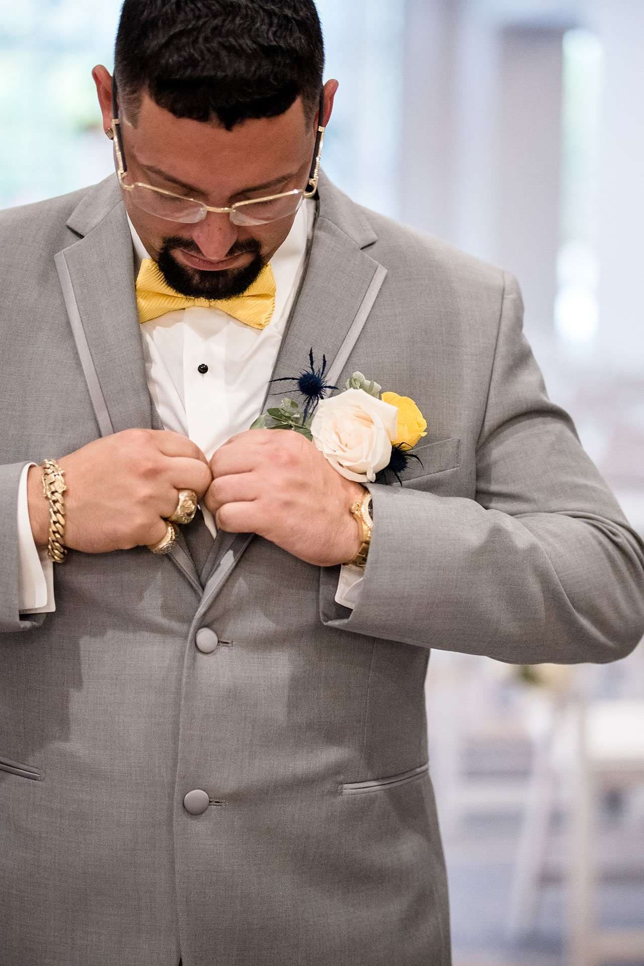 Groom in gray suit and yellow bow tie adjusting his jacket, with a white and yellow rose boutonniere