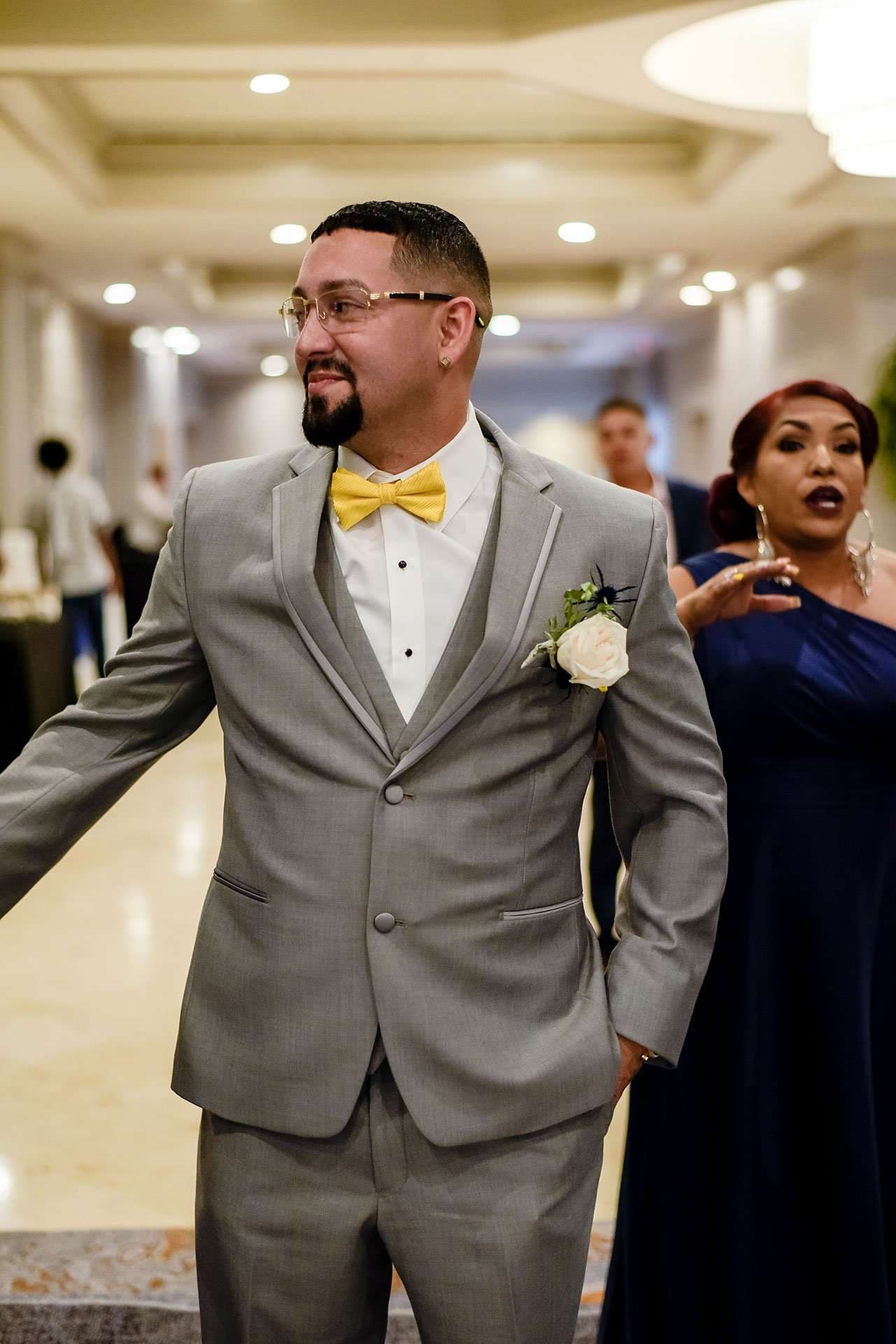 Groom in gray suit and yellow bow tie with a white boutonniere stands with arms out in a hotel hallway