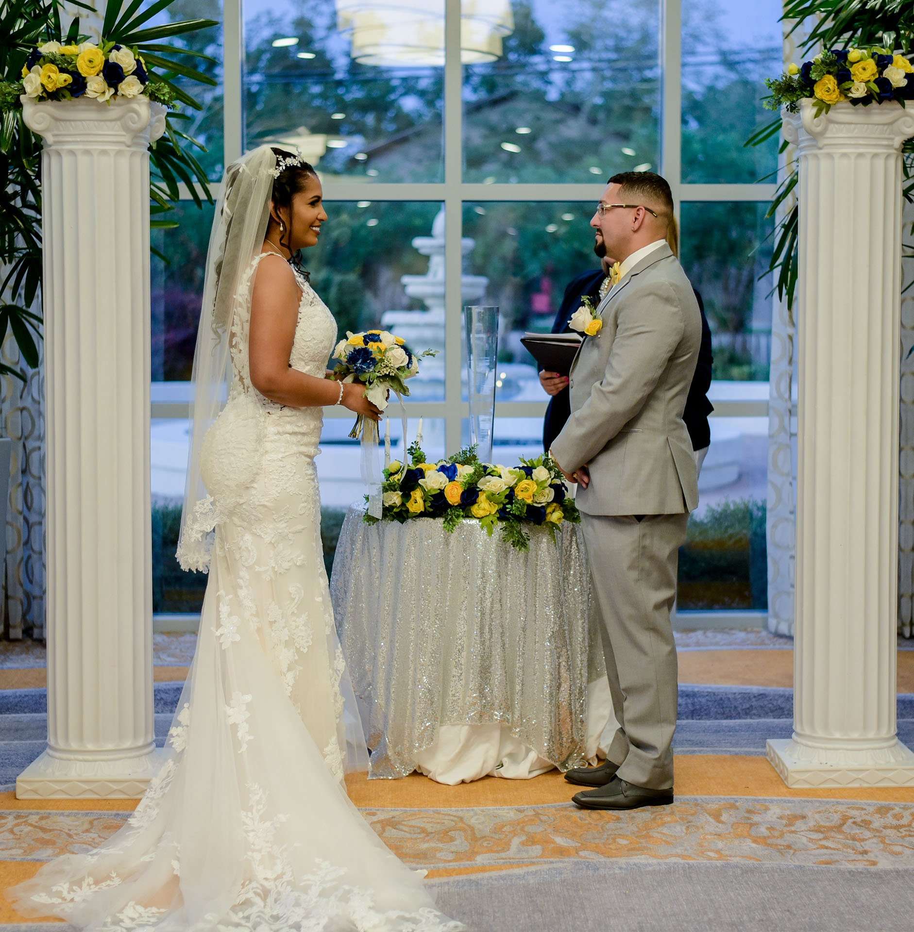 Bride in a lace gown and veil smiles at the groom in a gray suit during their ceremony between white columns